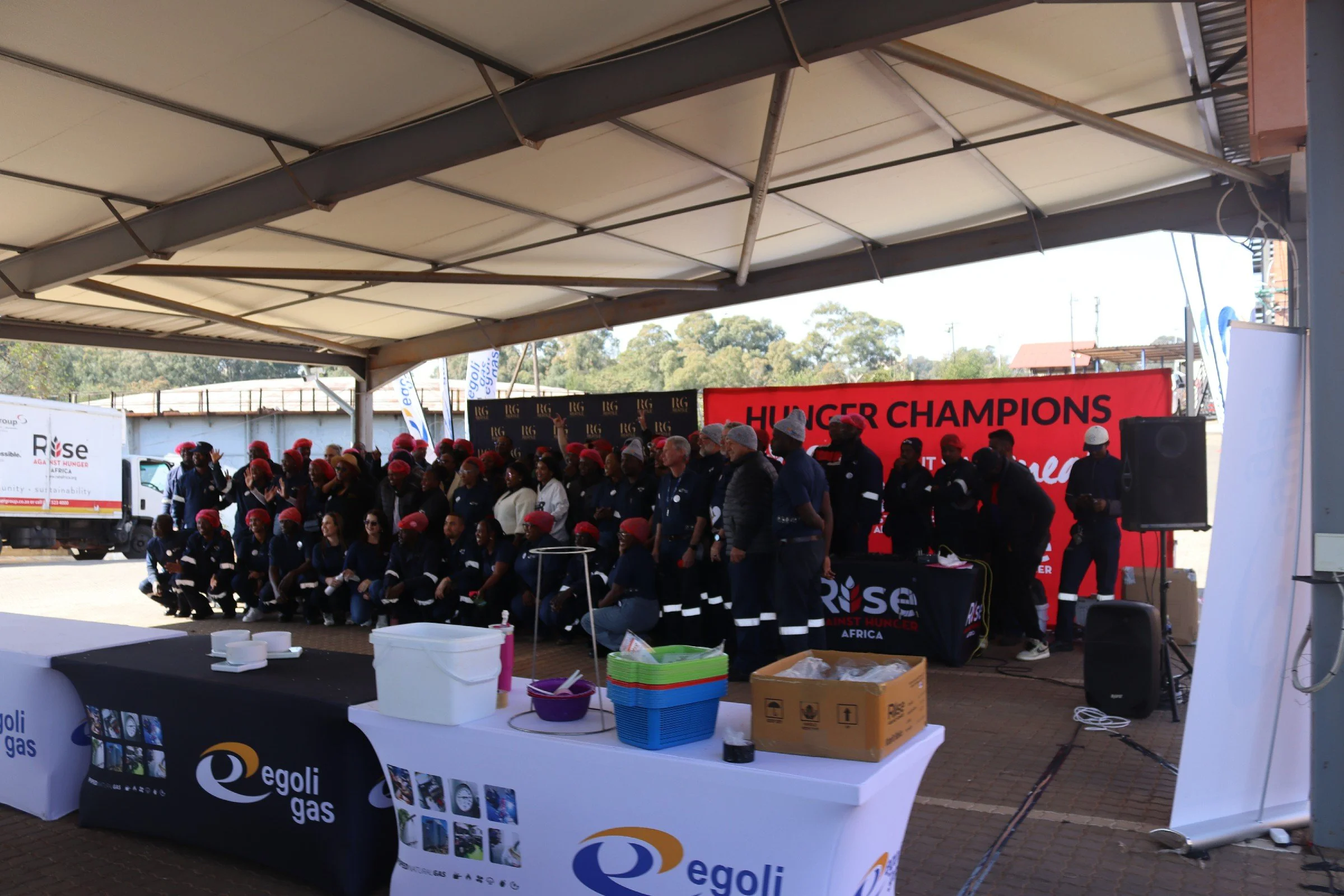 A large group of people wearing red hats and uniforms, gathered under a shelter at an outdoor event with banners reading 'Hunger Champions' and 'Rise Against Hunger.' There are tables with supplies in the foreground.