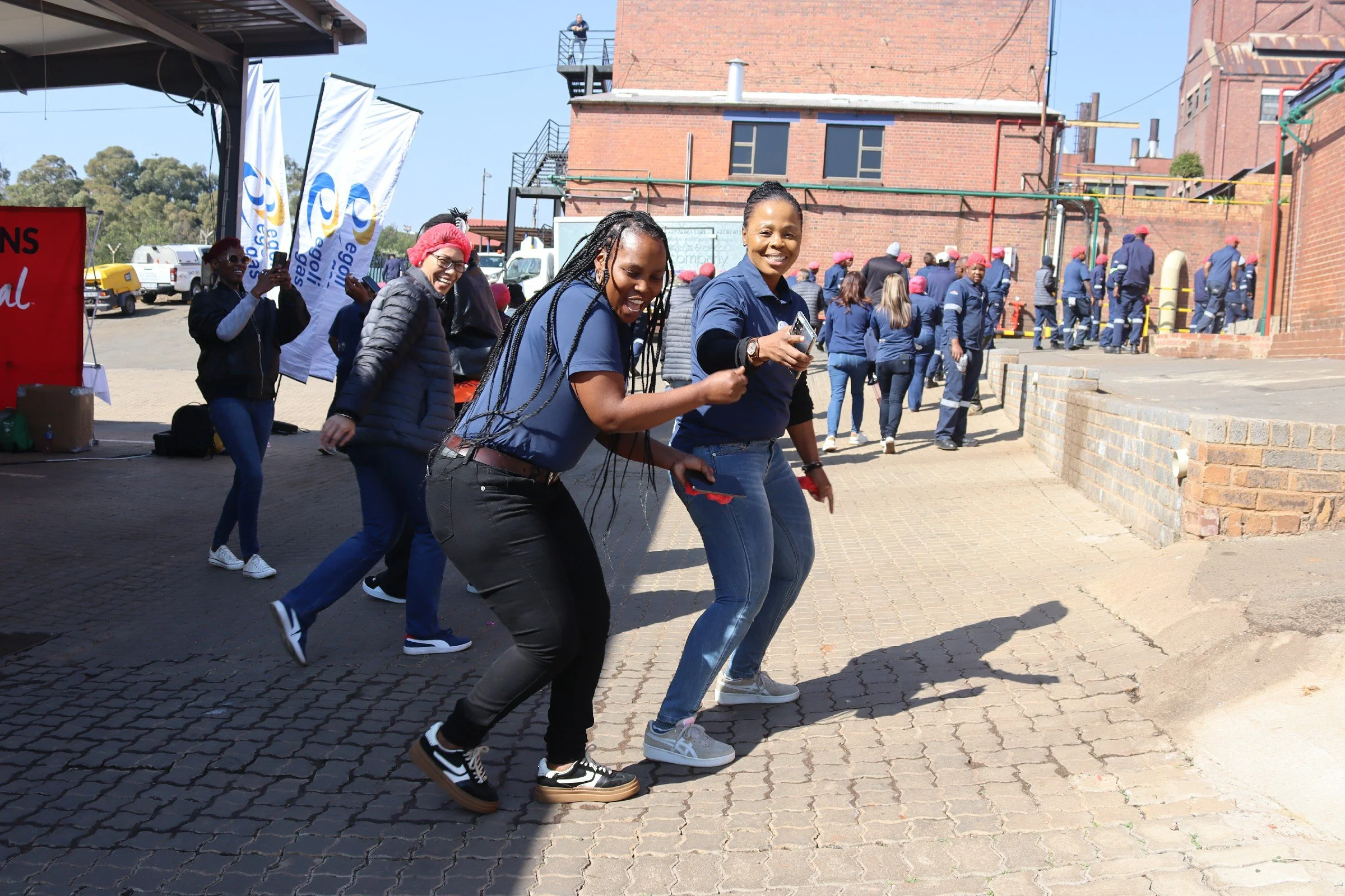 A group of women walking together outdoors, smiling and enjoying themselves, with a brick building, a row of people wearing uniforms, and flags in the background.