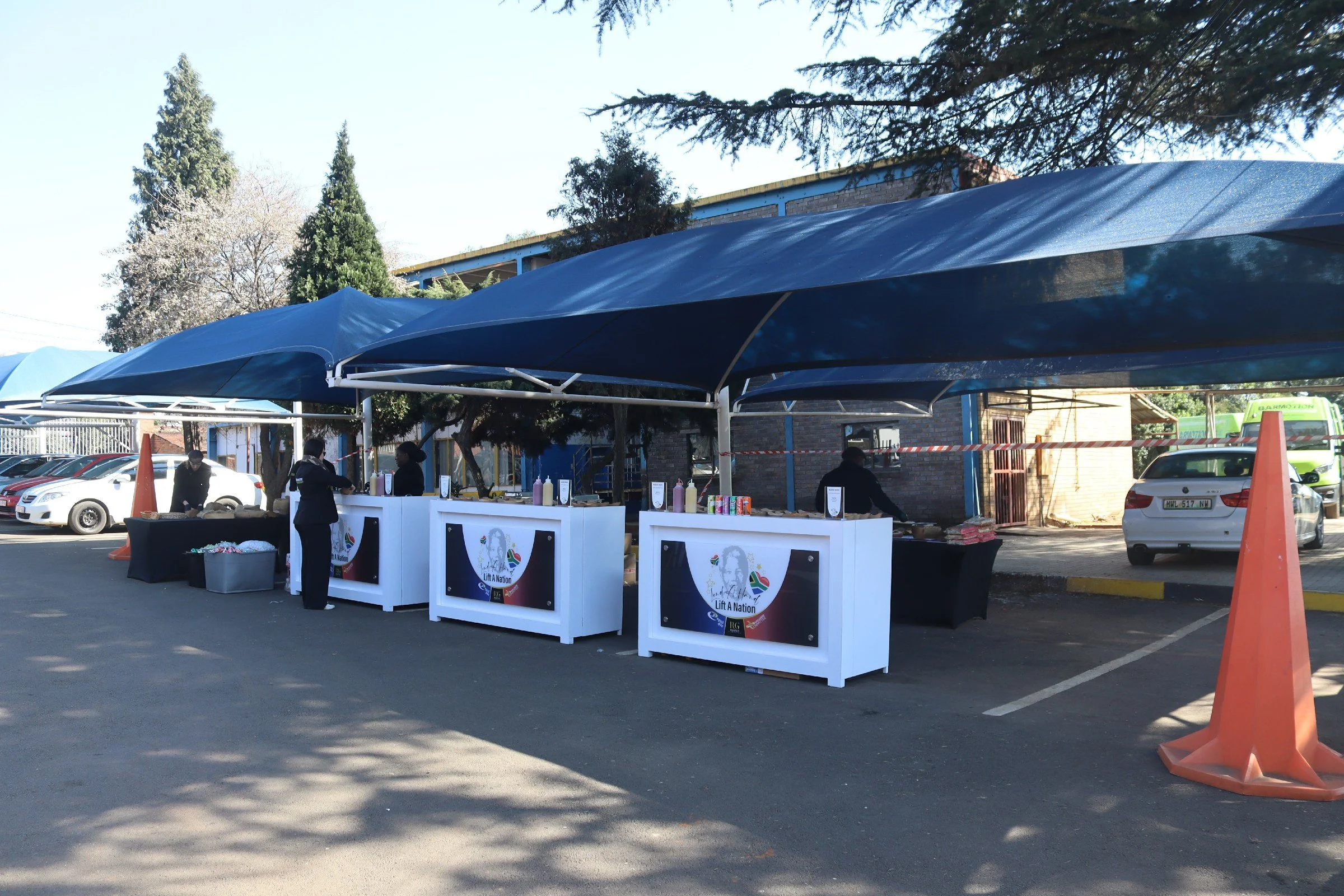 Outdoor event with multiple food stalls set up under blue canopy tents in a parking lot, with a few people working and cars parked nearby, trees in the background.