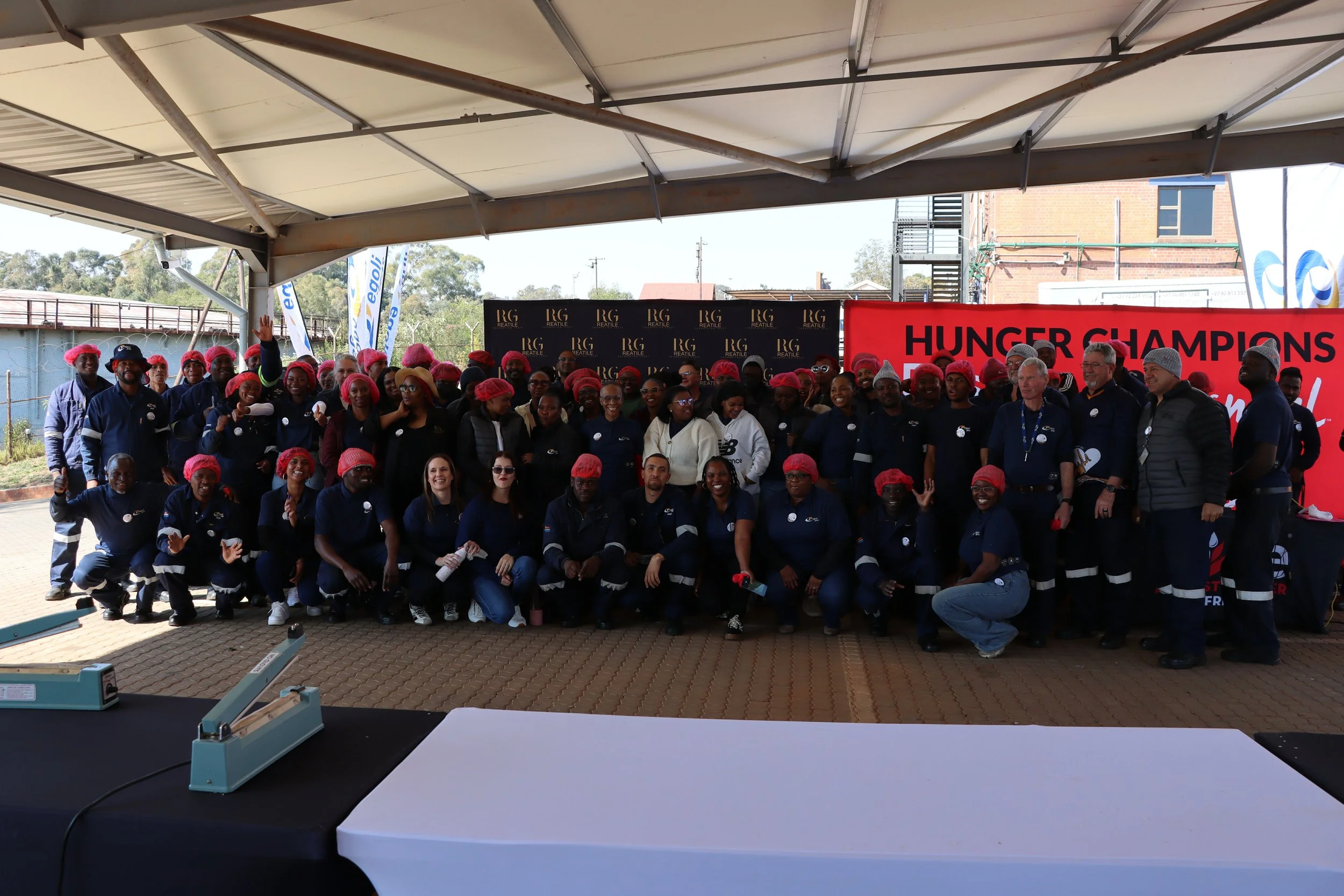 Group of people gathered outdoors under a large tent for a celebration or event, some wearing safety helmets, with a red banner that reads 'HUNGER CHAMPIONS' in the background.