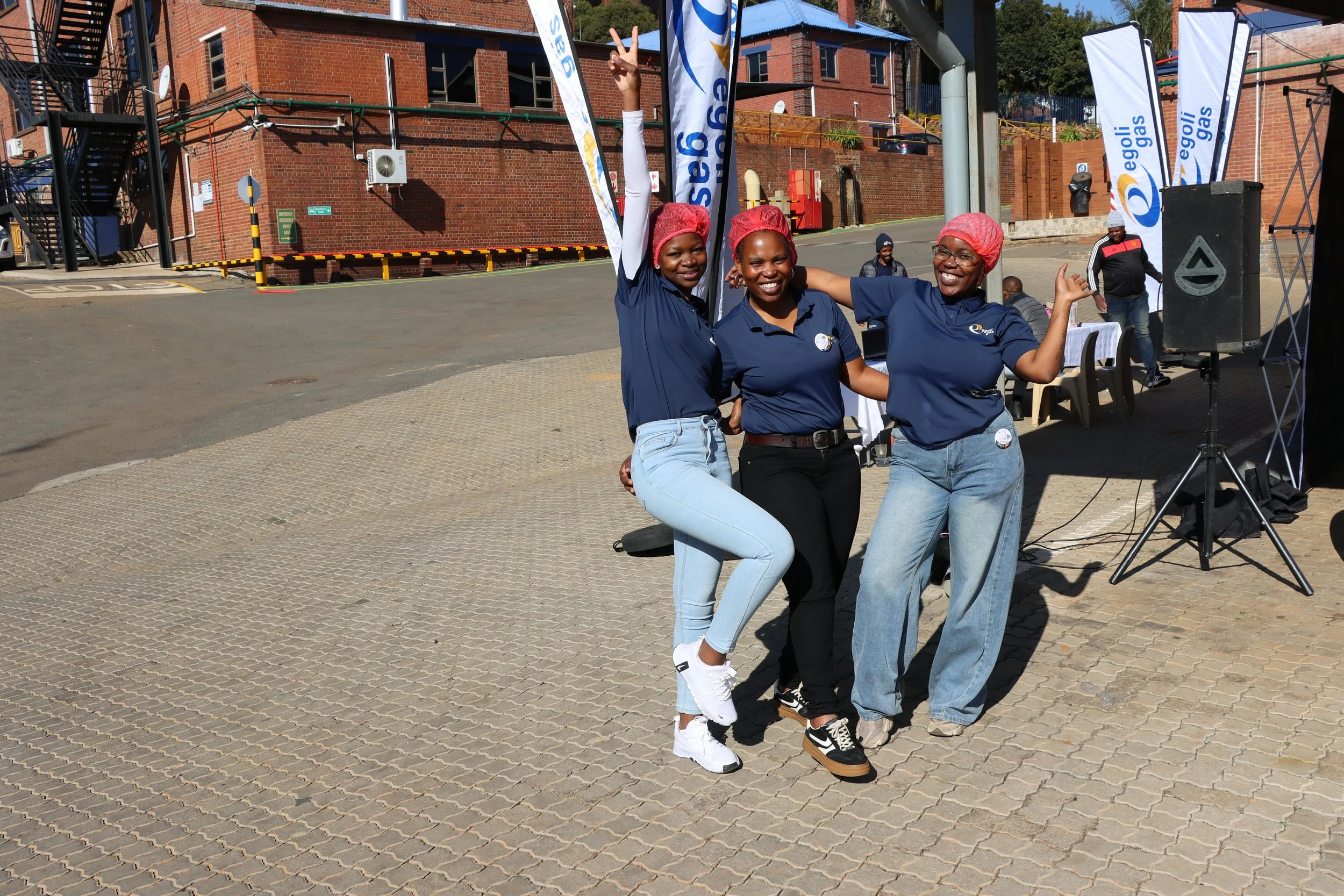 Three women wearing pink hairnets, blue shirts, and jeans posing together outdoors at a daytime event, with two giving peace signs and one making a hand gesture, in front of banners and a brick building.