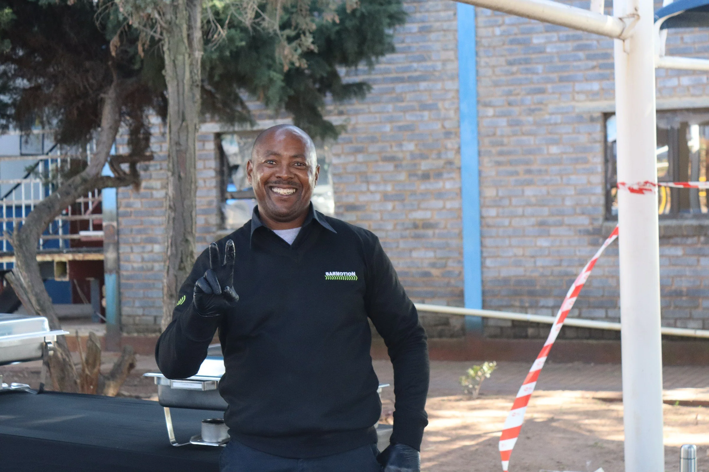 A smiling man wearing a black long sleeve shirt and black gloves standing outdoors next to a car, making a peace sign with his right hand. In the background, there is a brick building, a tree, and some construction tape.