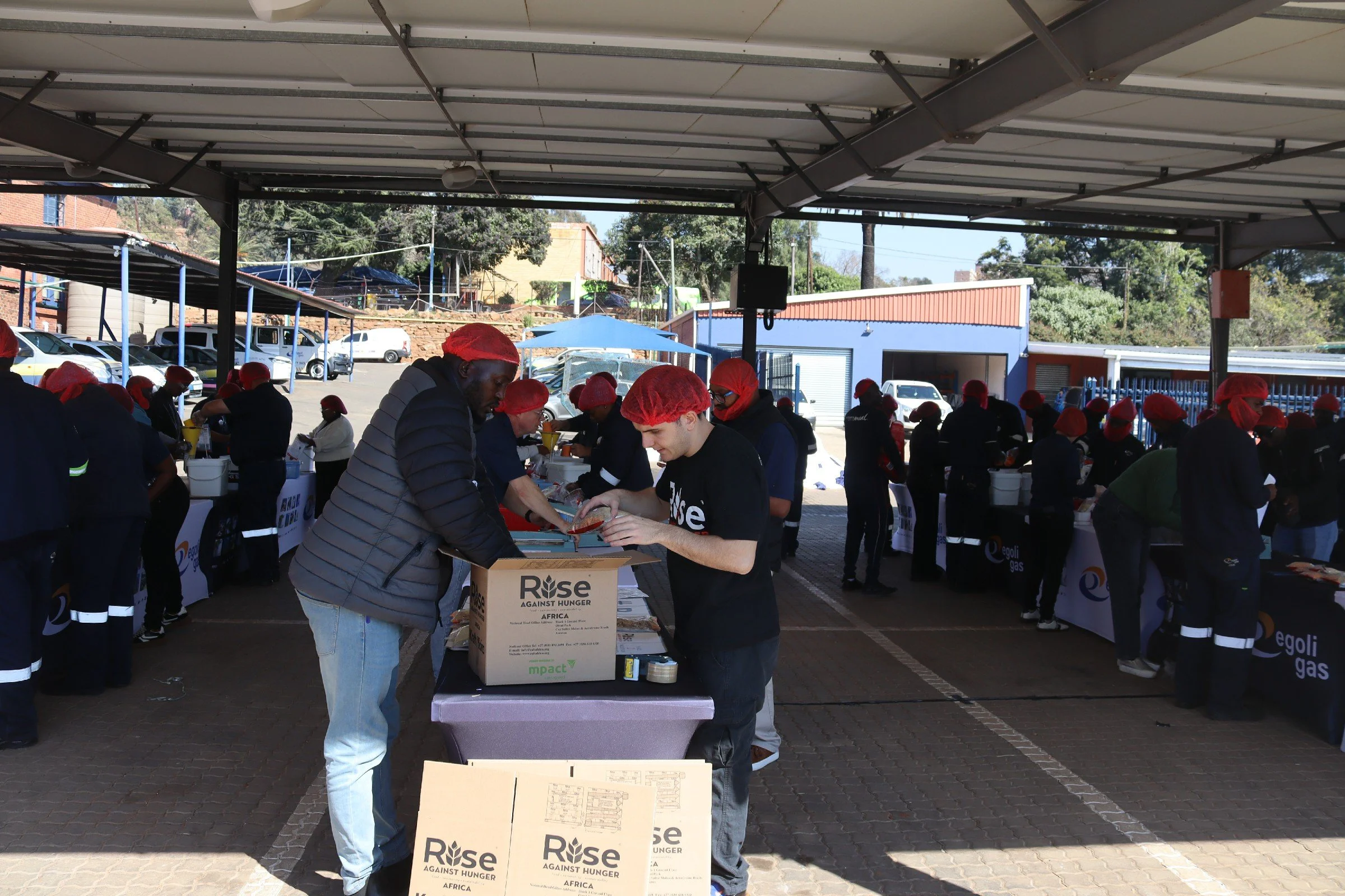 People wearing red hairnets standing under a carport, organizing boxes for a food distribution event, with cars parked nearby and a blue building in the background.