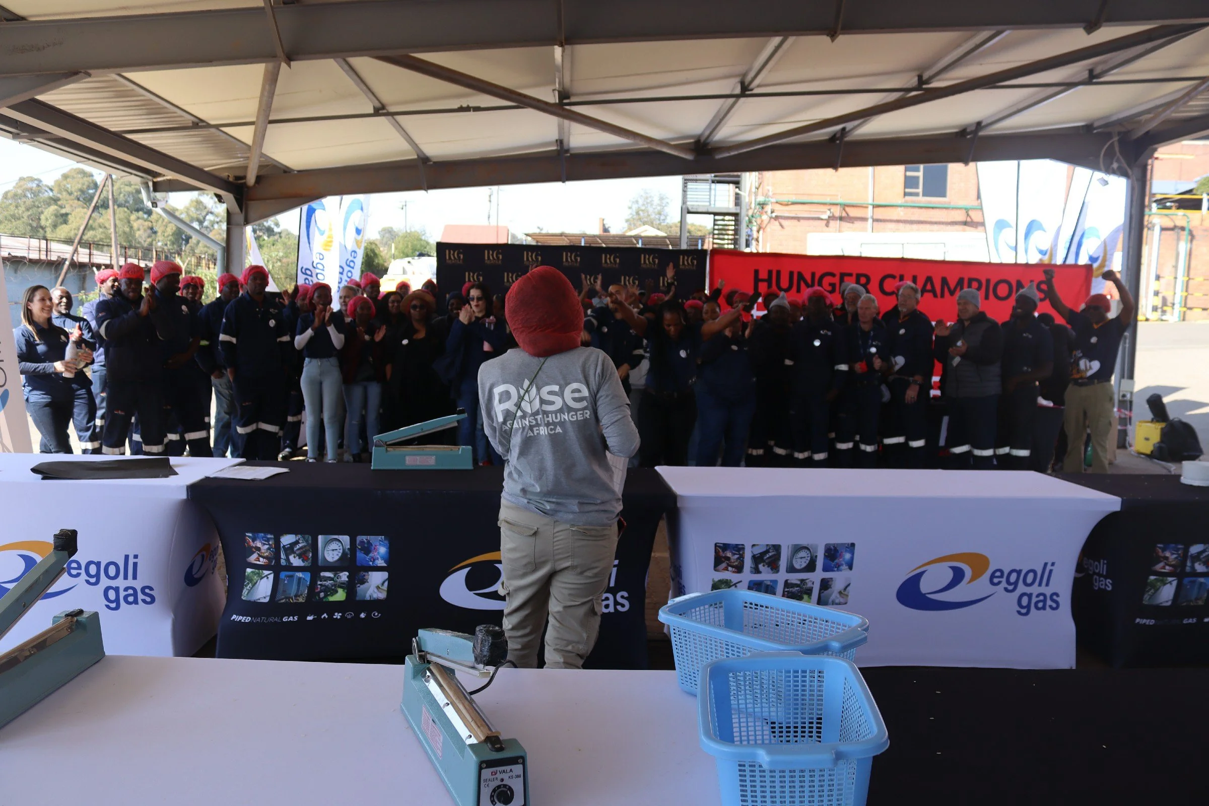 Group of people wearing red head coverings and dark uniforms under a canopy, some clapping, with a banner that reads "HUNGER CHAMPIONS" in the background, and a person in a gray "Rise Against Hunger Africa" shirt in the foreground.
