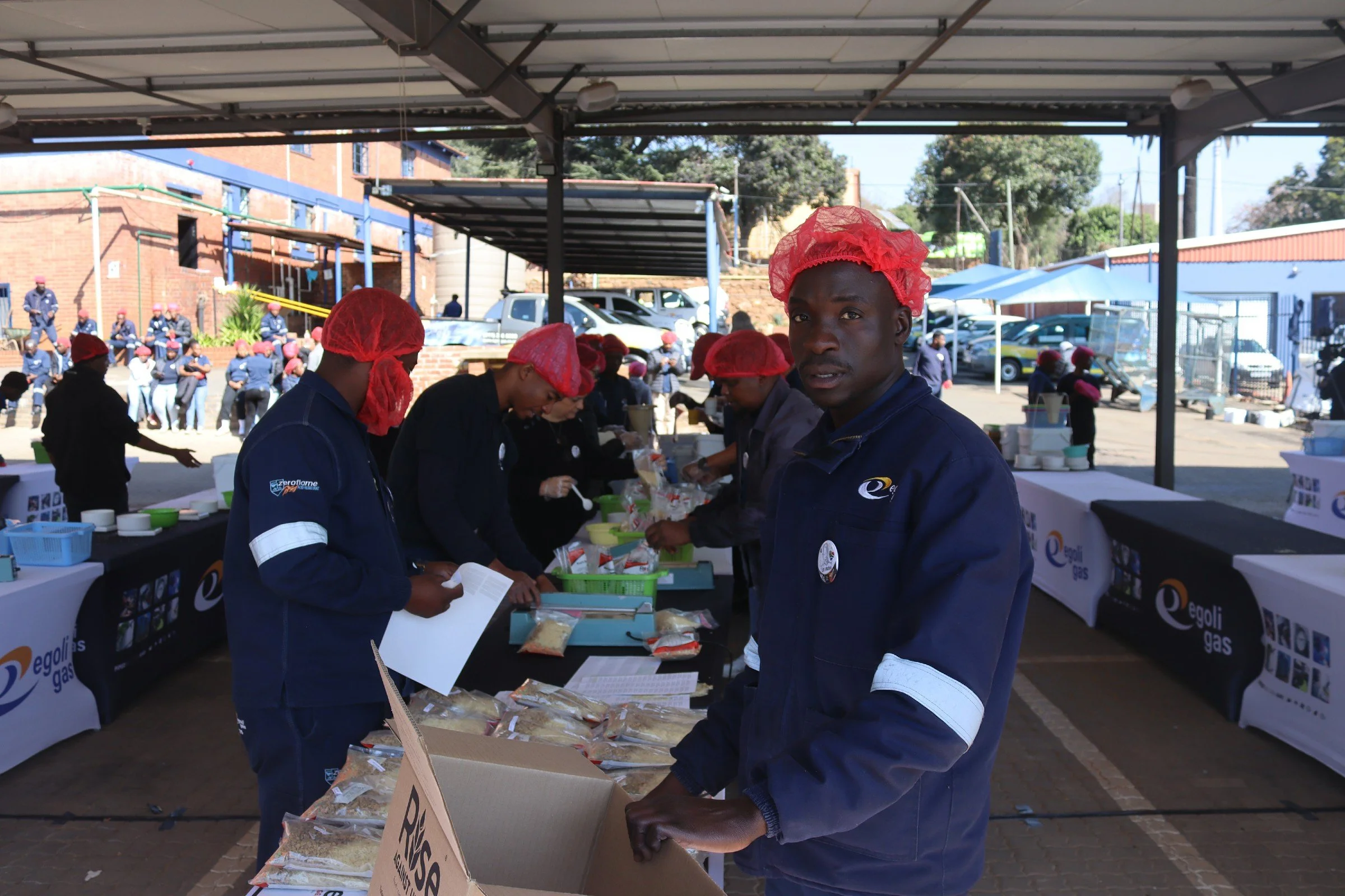 People wearing red hairnets and blue uniforms working at outdoor food distribution table, with onlookers seated in the background.