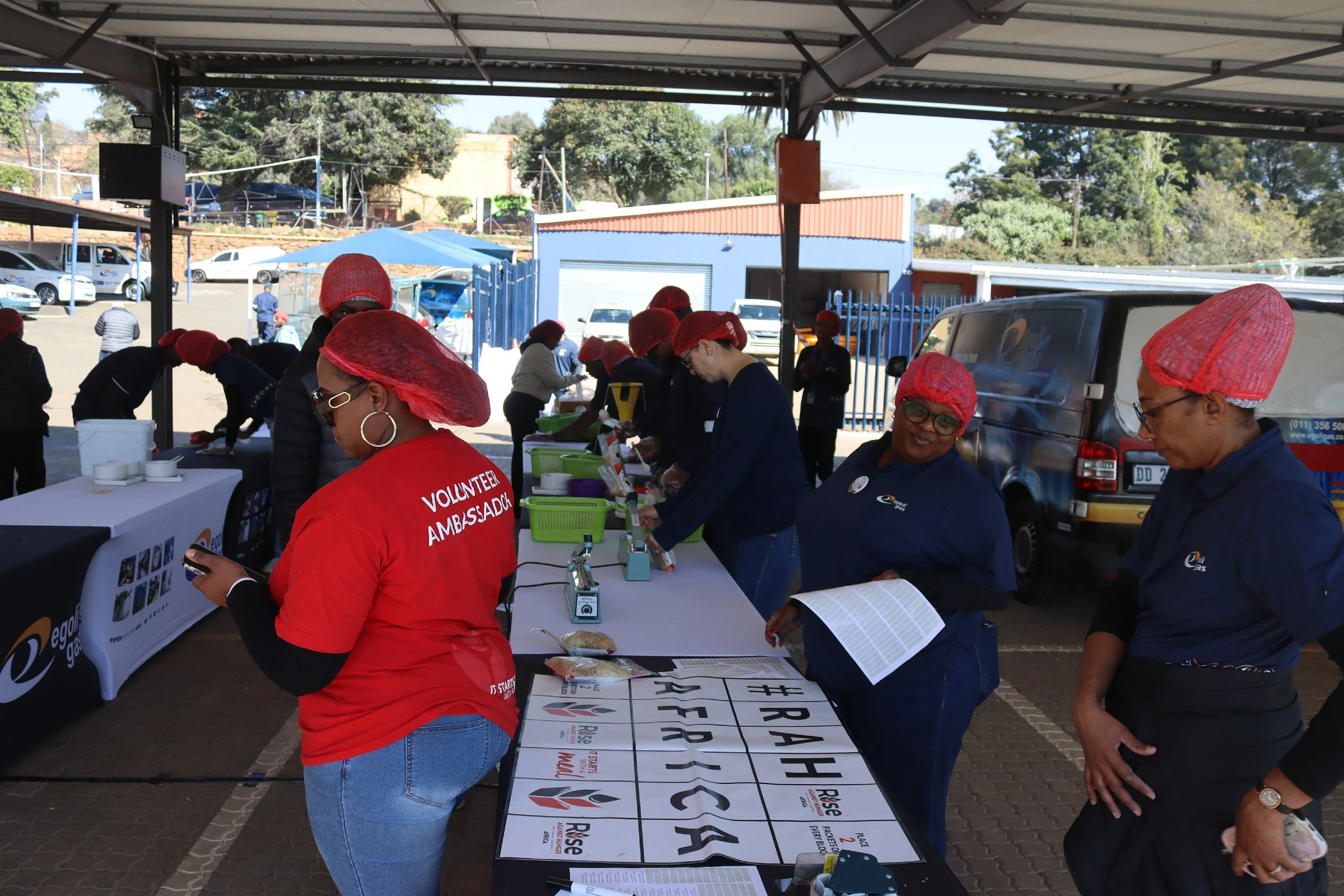 Group of people at an outdoor event, wearing red hairnets and working at tables, with signs and equipment, under a shaded area.