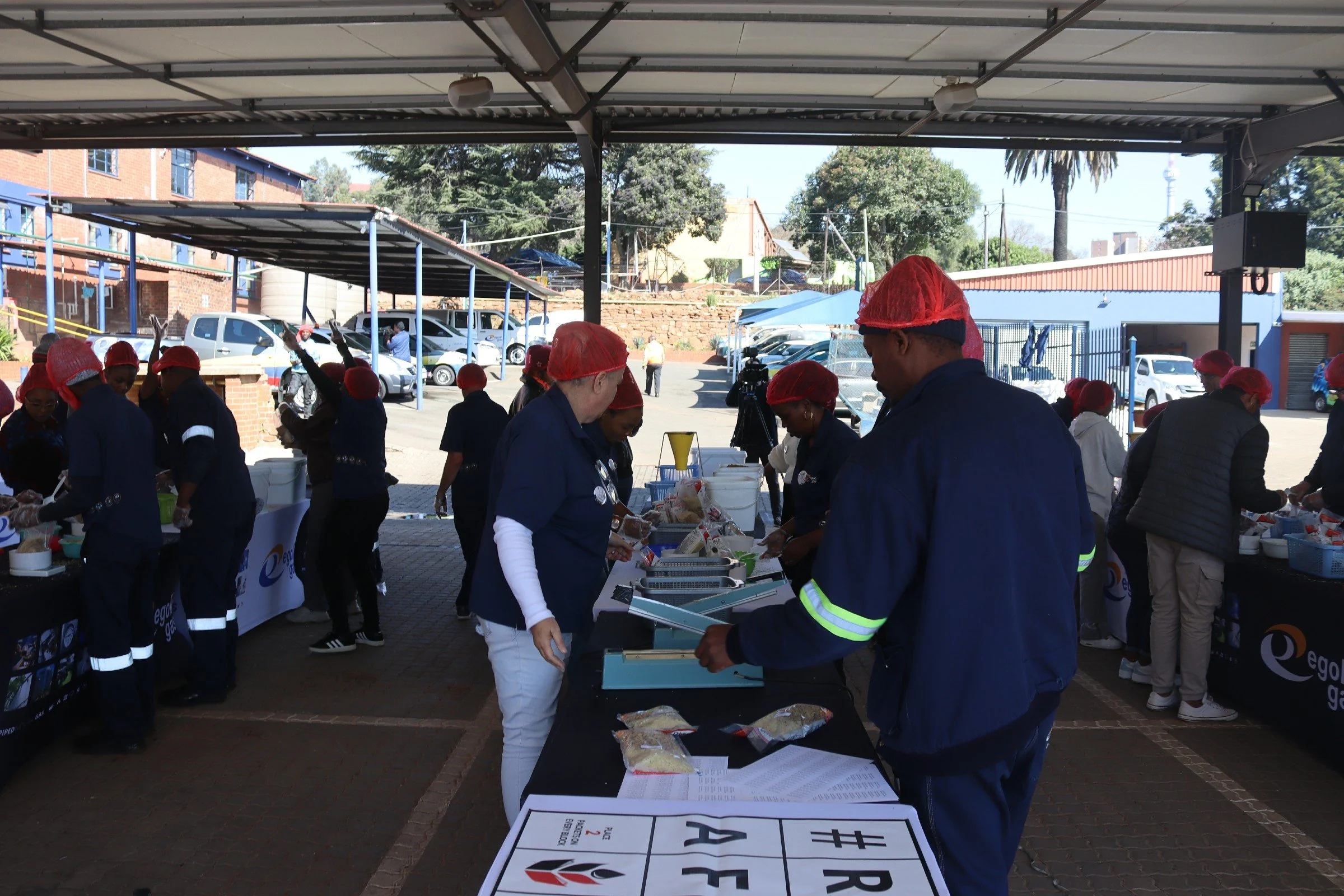 People wearing red hairnets and navy uniforms gather around tables under a covered structure, participating in an outdoor food distribution or cooking event. Several cars are parked in the background, along with trees and buildings.