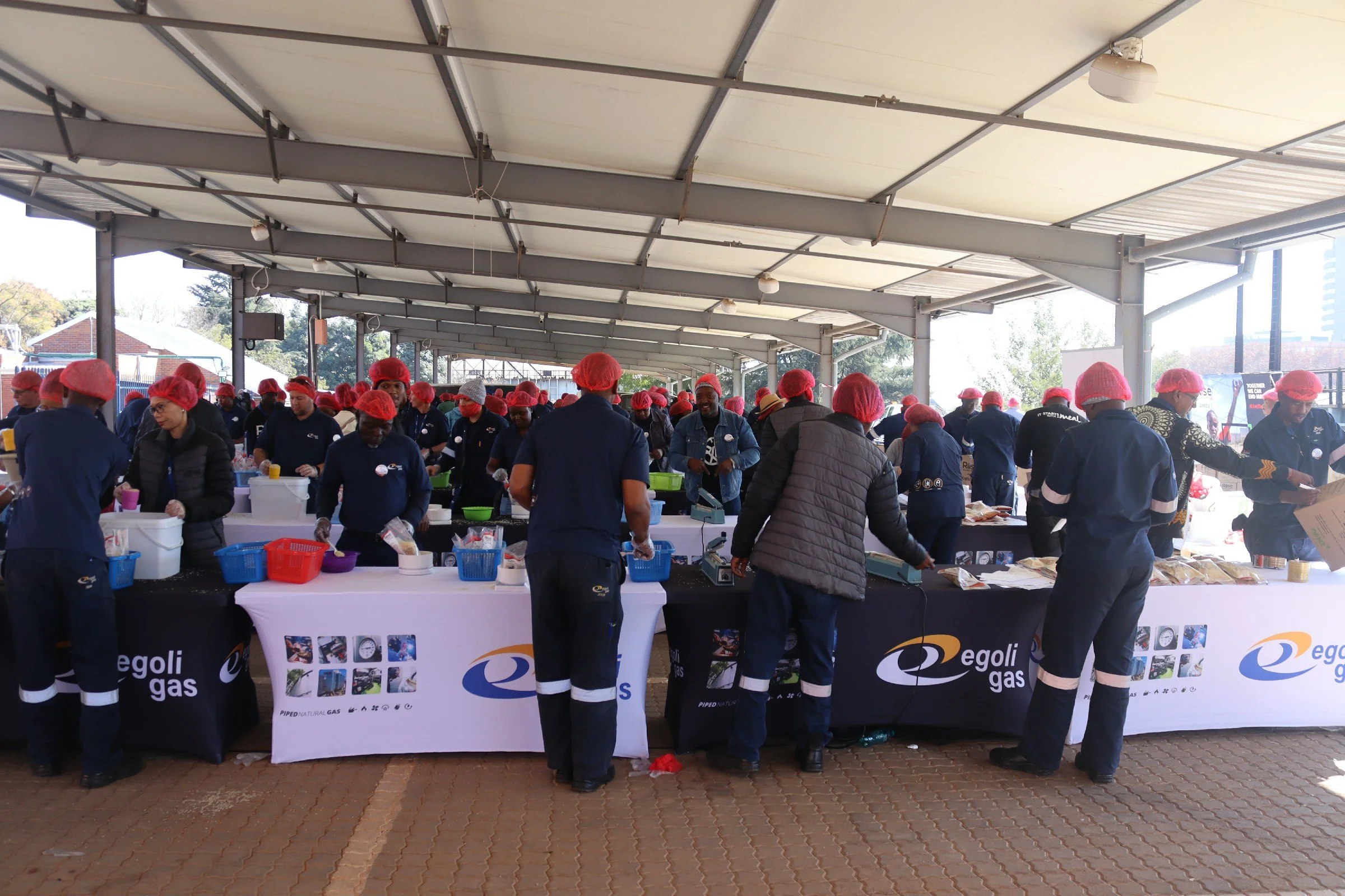 People working at a food stall under a large outdoor awning. They are wearing blue uniforms and pink hair nets, preparing and serving food. The stall has a table with various supplies and a banner with the text 'egoli gas' and images of gas-related i