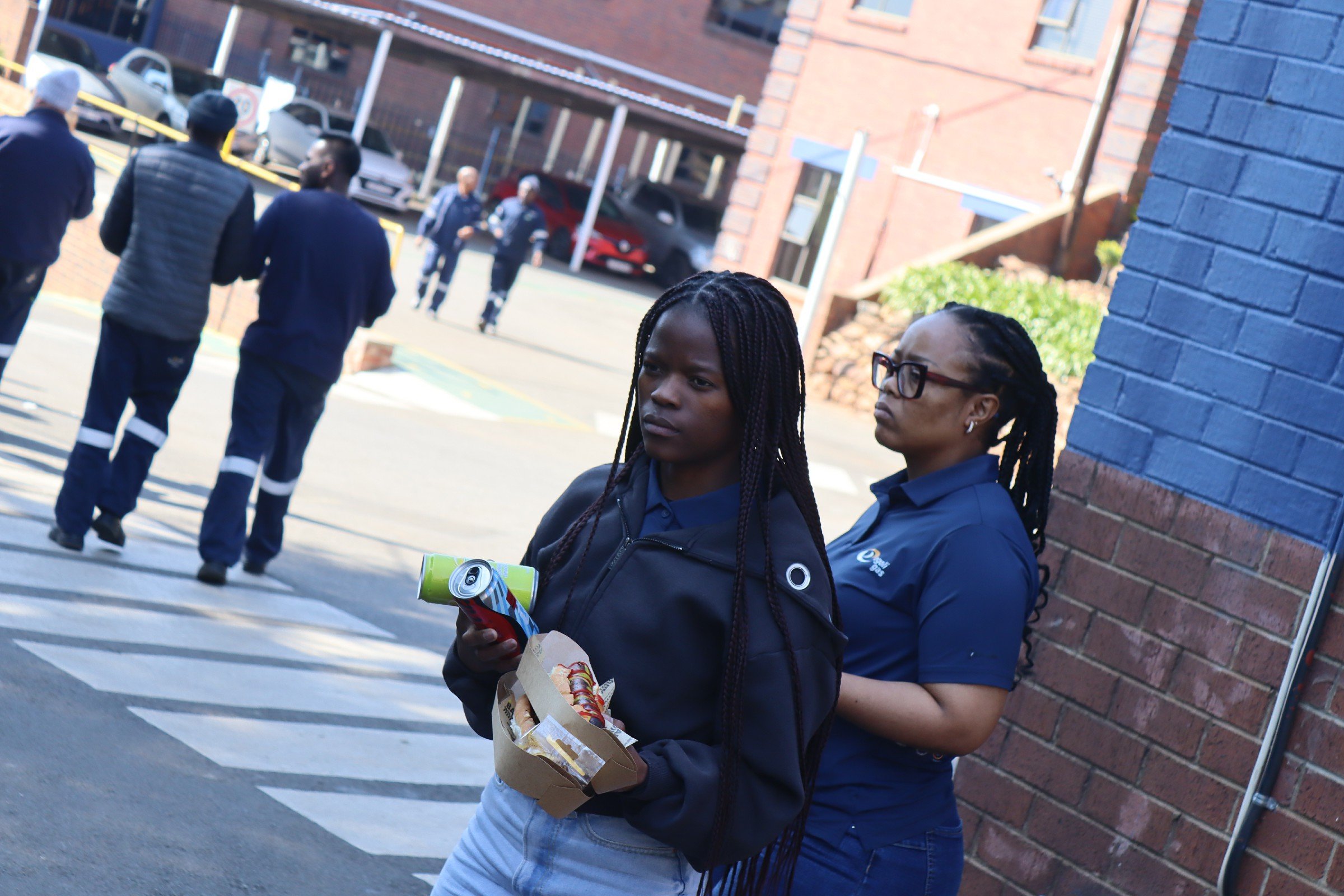 Two women standing against a brick wall, one holding a snack and a drink, the other wearing glasses, with people walking in the background outside a multi-story parking garage.