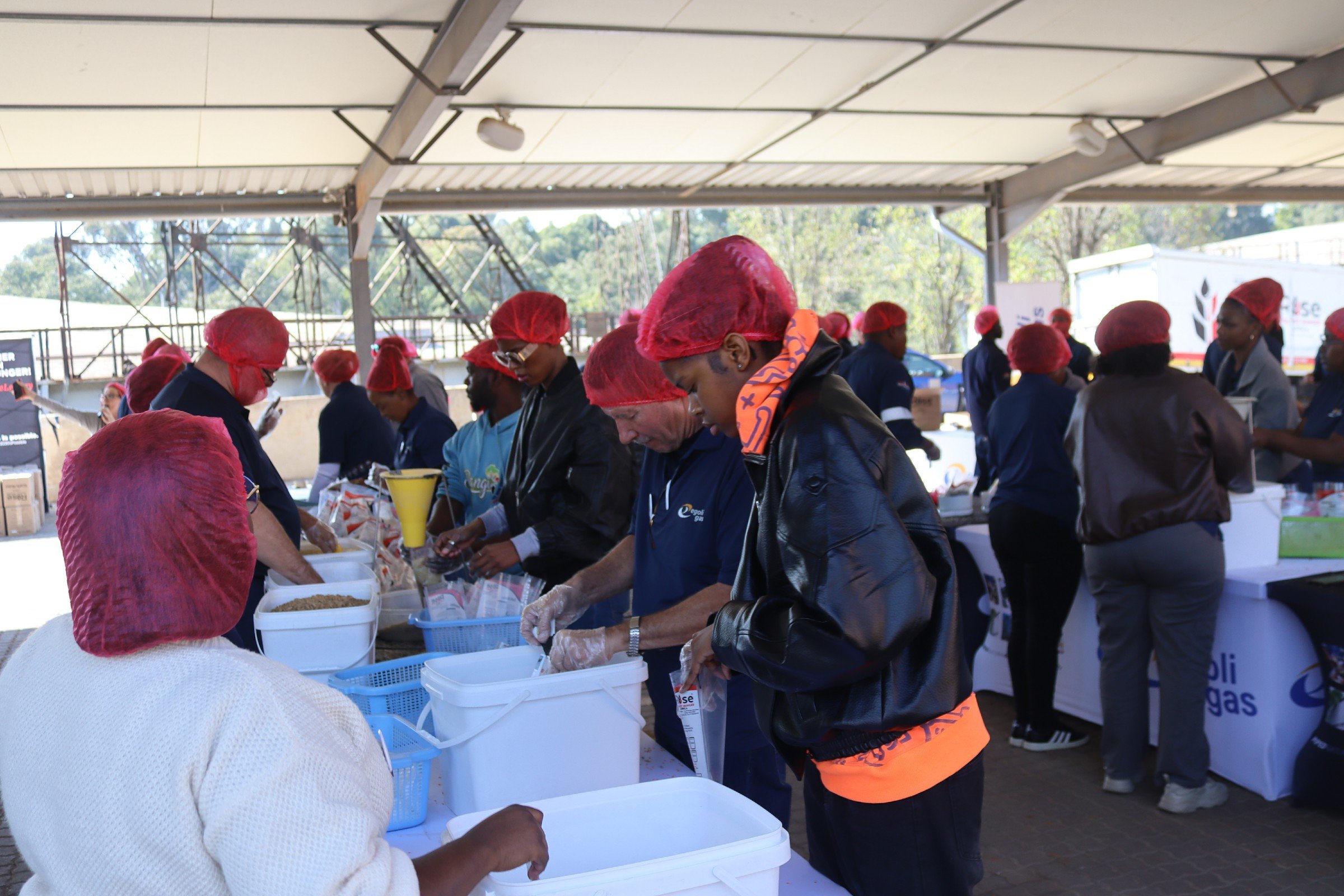 Several people wearing red hairnets and gloves working together at a table, possibly preparing food or packaging items at an outdoor event or food bank.