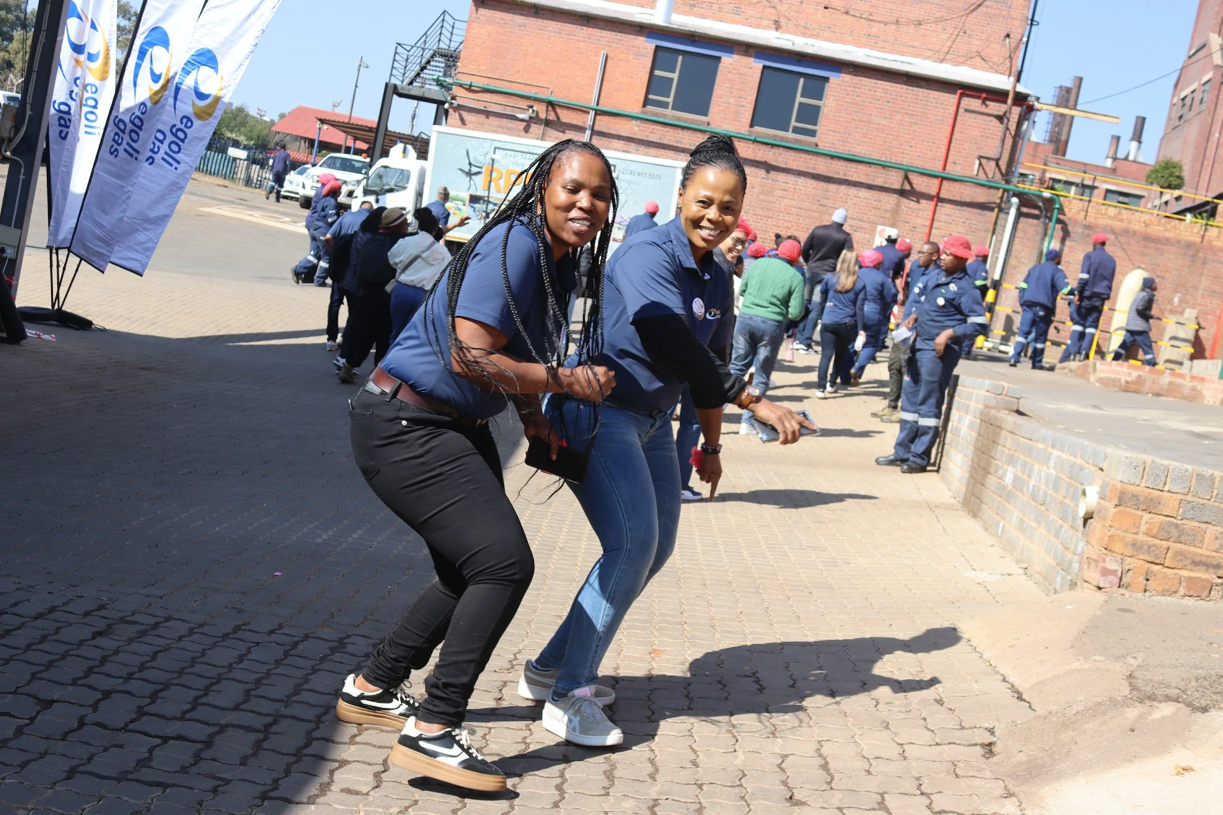 Two women smiling and skipping on a cobblestone street during an outdoor event, with a crowd and brick buildings in the background.
