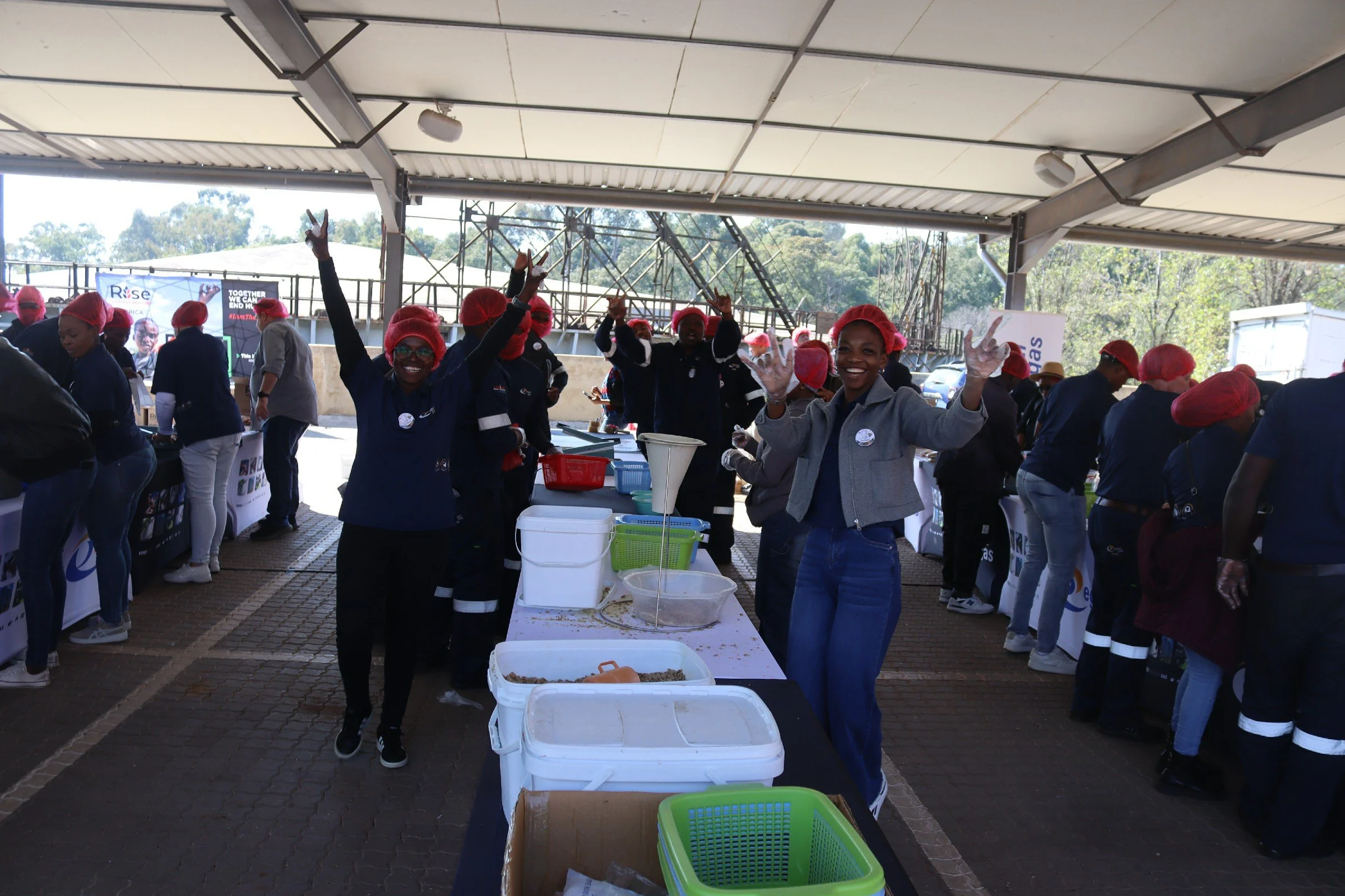 Group of women wearing red hairnets and gloves, smiling and making peace signs, standing behind a table with containers of food at an outdoor event under a large tent.