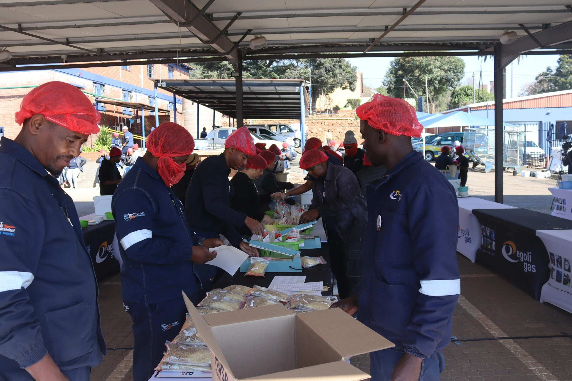 People wearing blue uniforms and red hair nets at an outdoor event, preparing or serving food at tables under a canopy with parked cars and trees in the background.