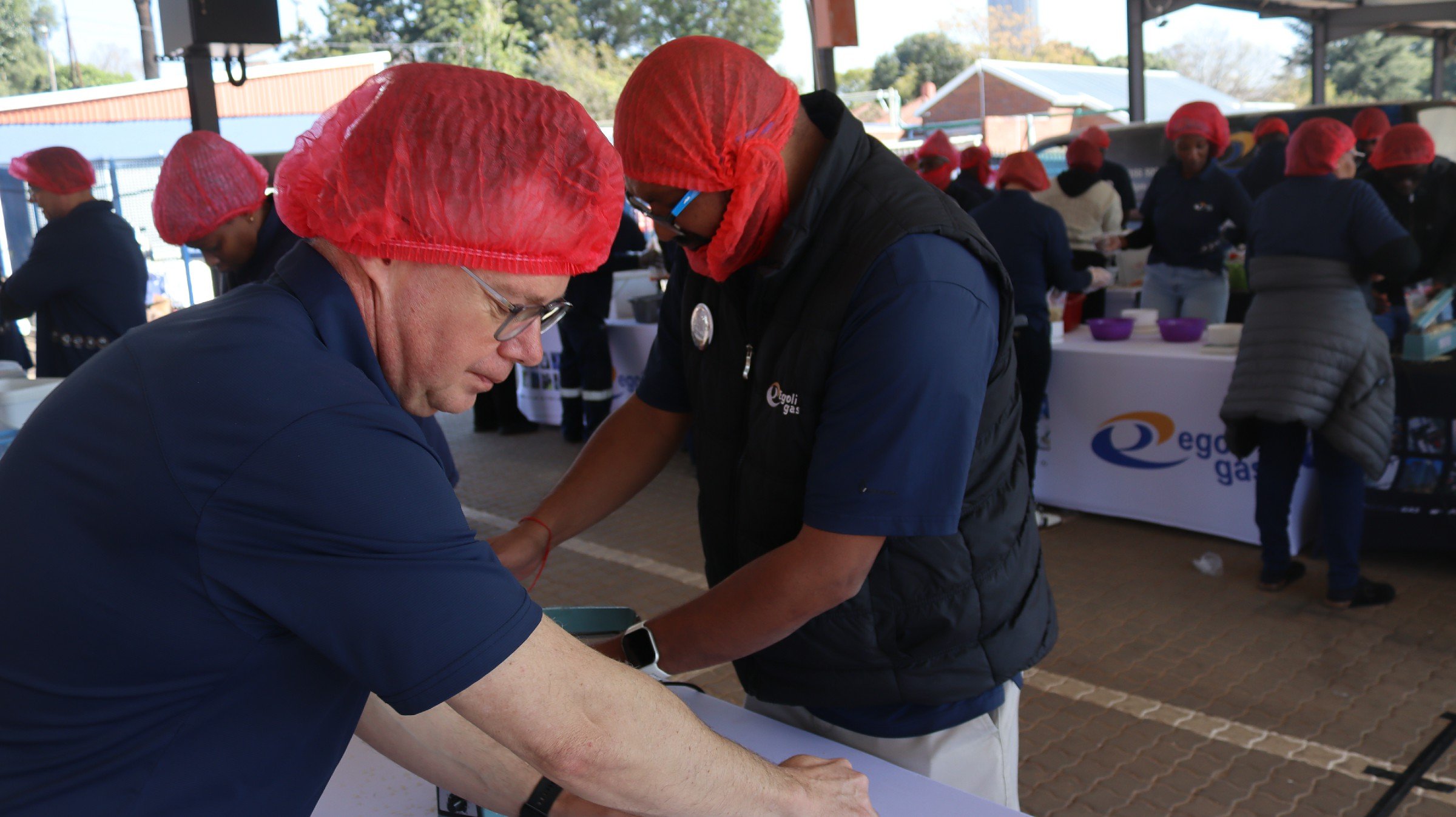 People wearing red hair caps working together at a table during an outdoor event, with other participants and booths in the background.