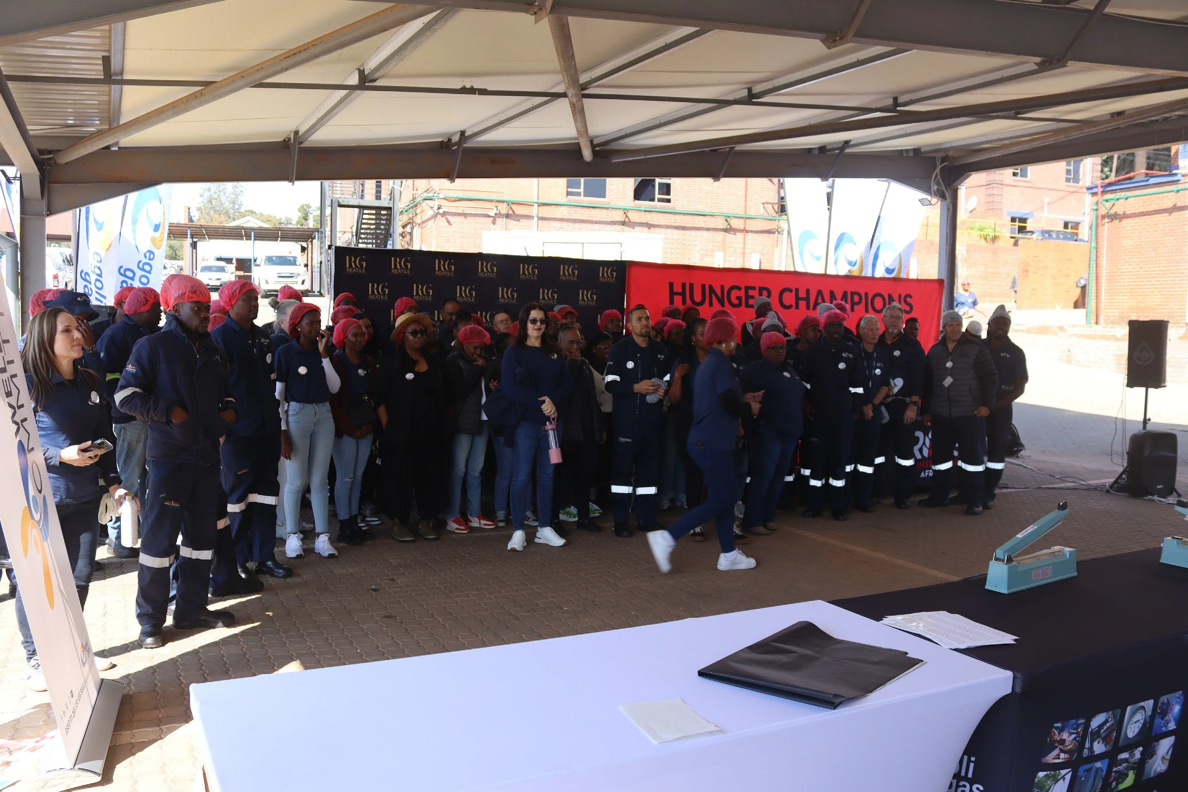 Group of people, mostly women, wearing red hairnets and dark uniforms, standing under a large canopy at a Hunger Champions event.
