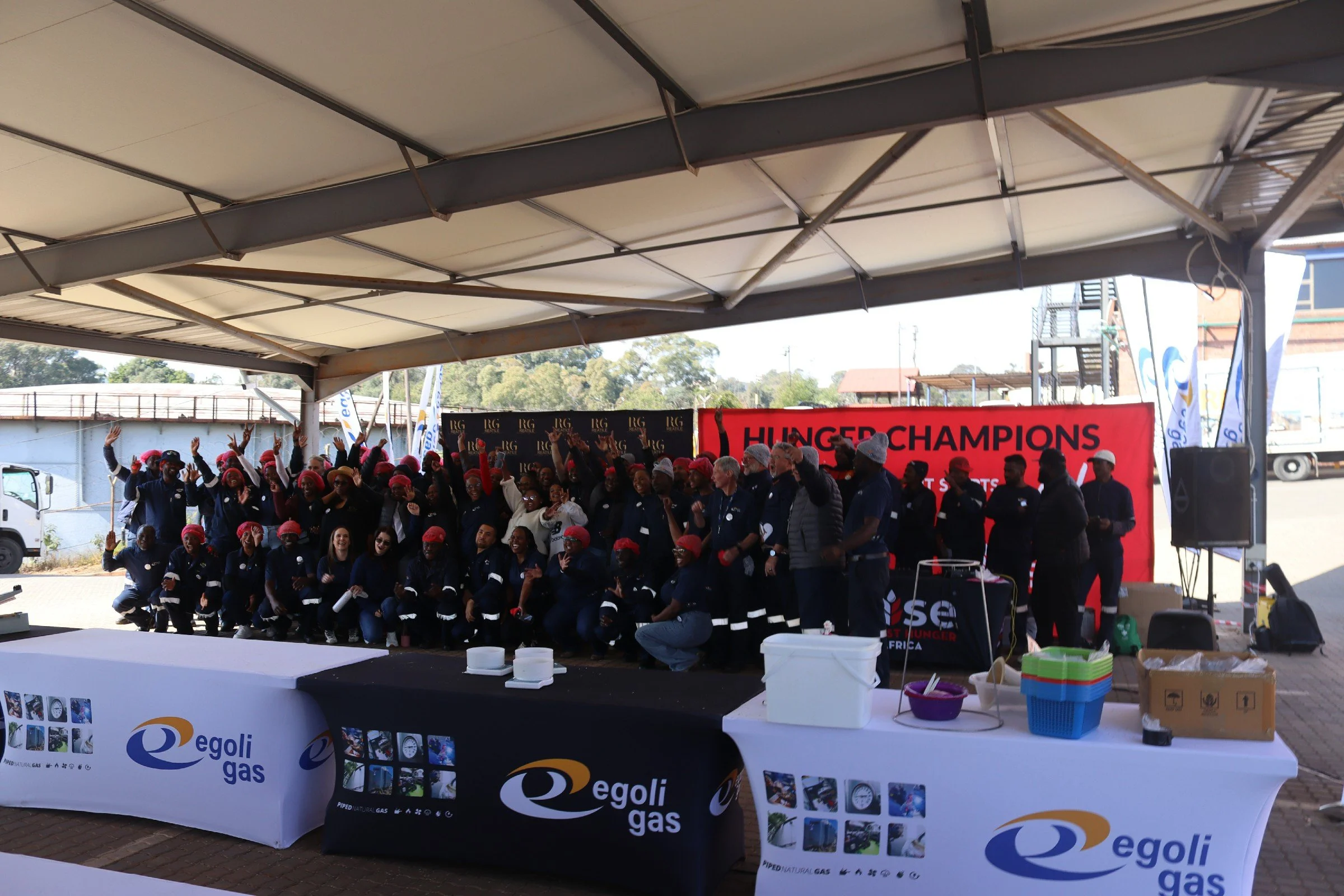 Group of people participating in a community event or rally under a large canopy, with banners and tables displaying Egoli gas branding and promotional materials.