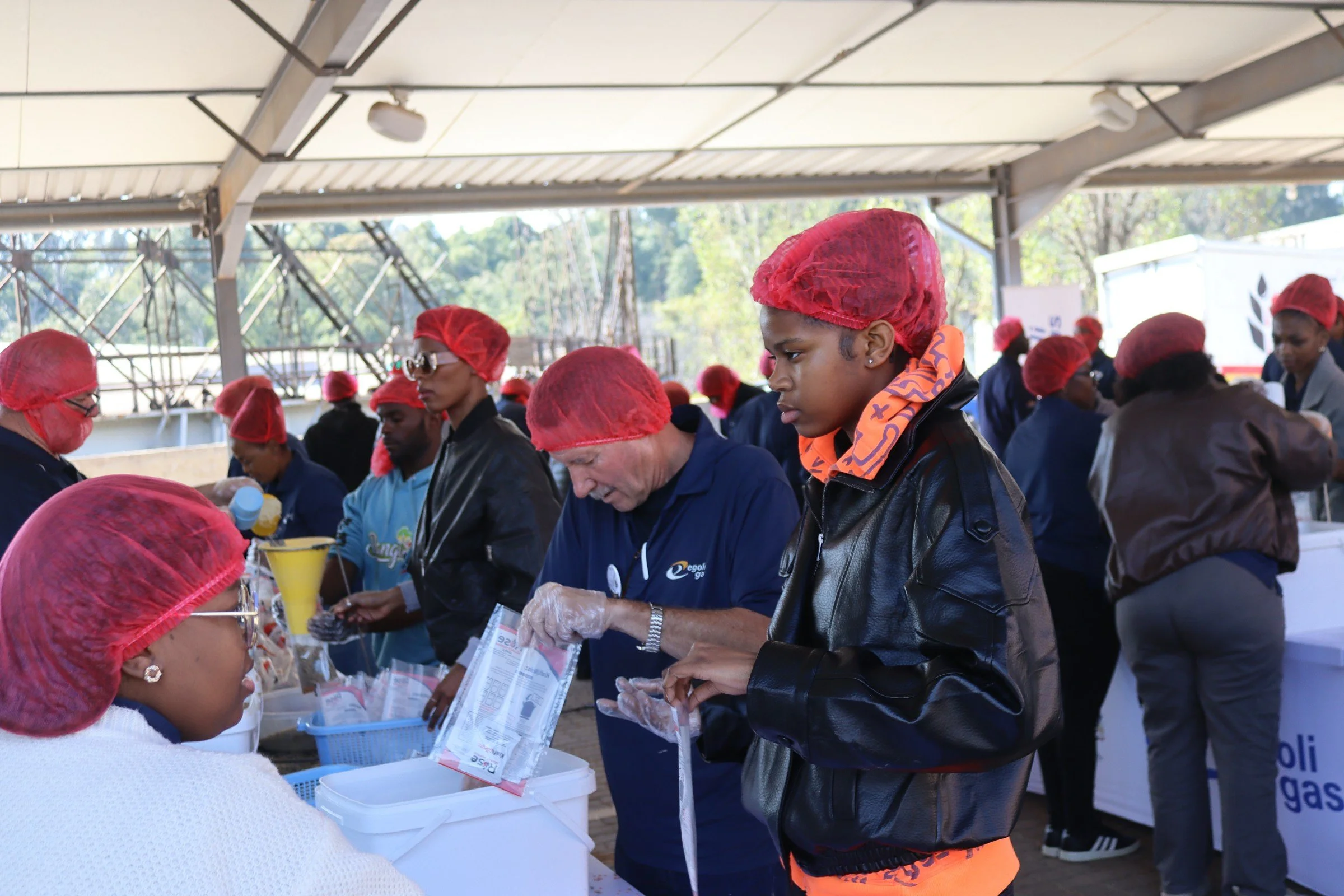 People working at a food distribution event, wearing red hair covers, with some holding papers and bags, under a large outdoor shelter with trees in the background.