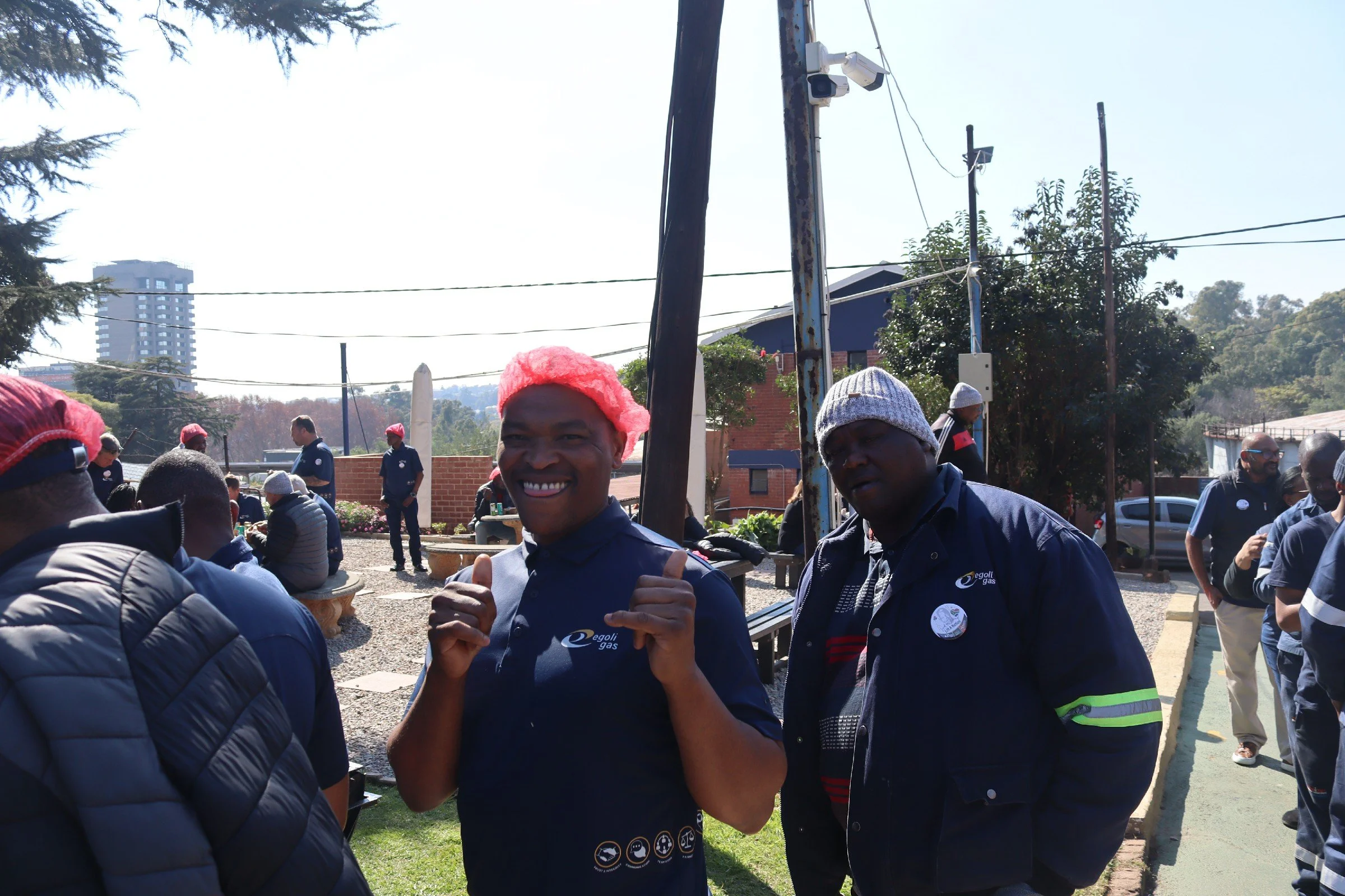 A group of people outdoors participating in an event, with a man in the center giving a thumbs-up, wearing a dark shirt and a red hat. Others are gathered around, some wearing winter hats and jackets, with trees, power lines, and a tall building in t