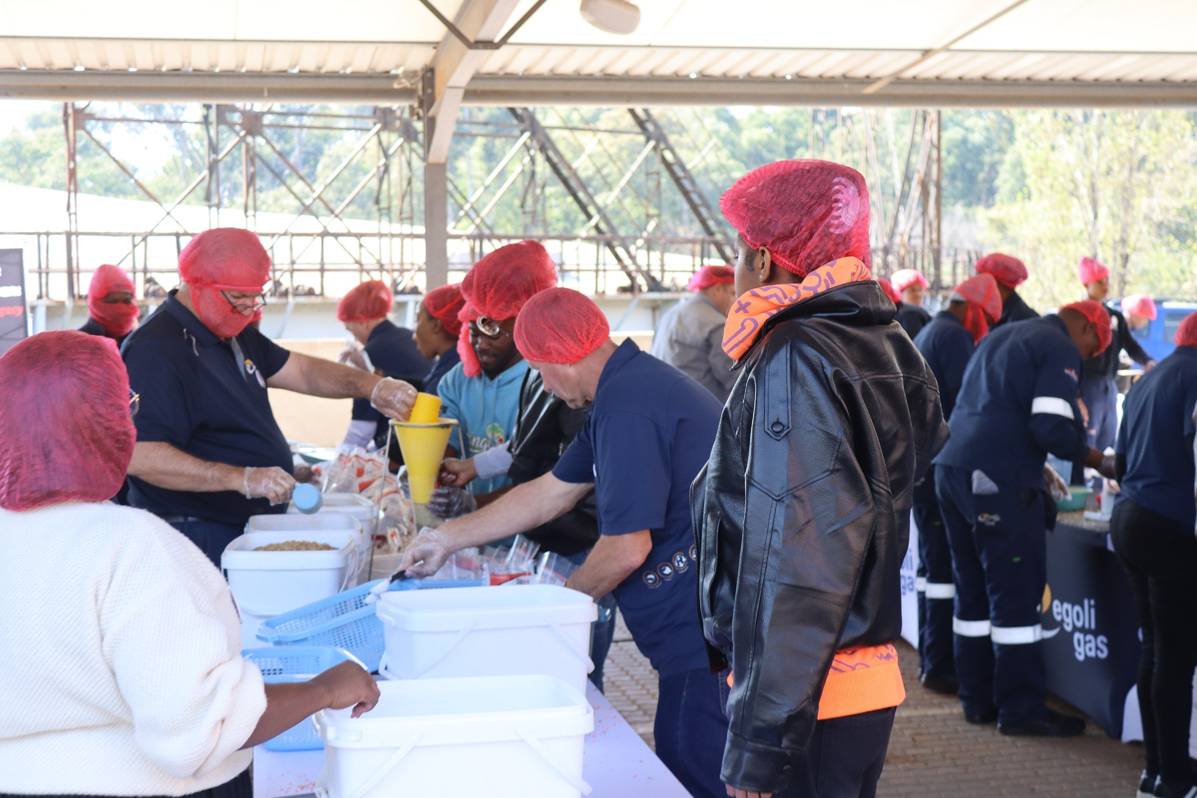People wearing red hairnets and gloves working at a long table under a metal canopy, preparing food or packages outdoors on a sunny day.