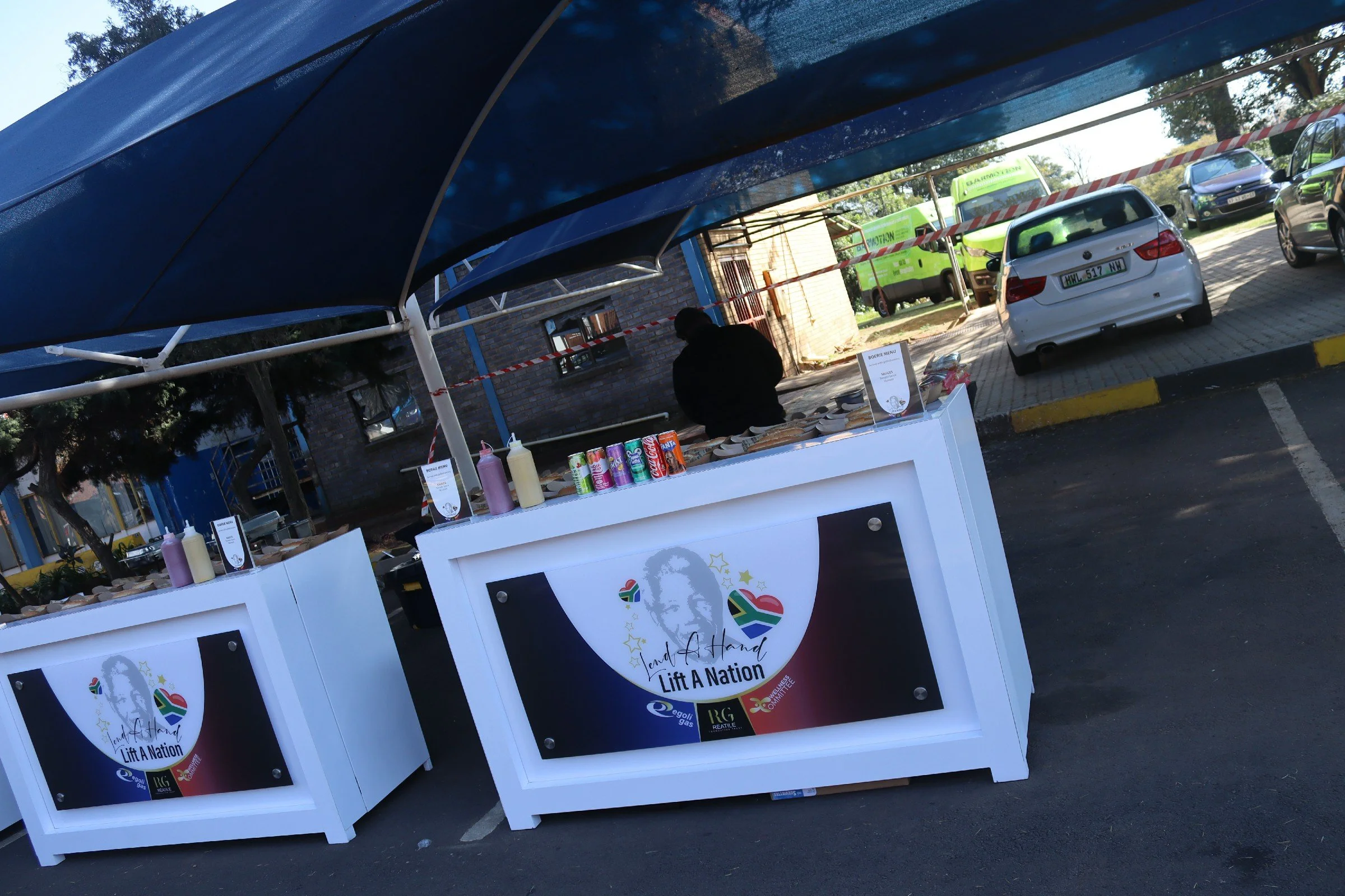 Outdoor stall with a colorful logo promoting lifting the nation, featuring a face with stars and South African flag elements, set under a blue canopy.