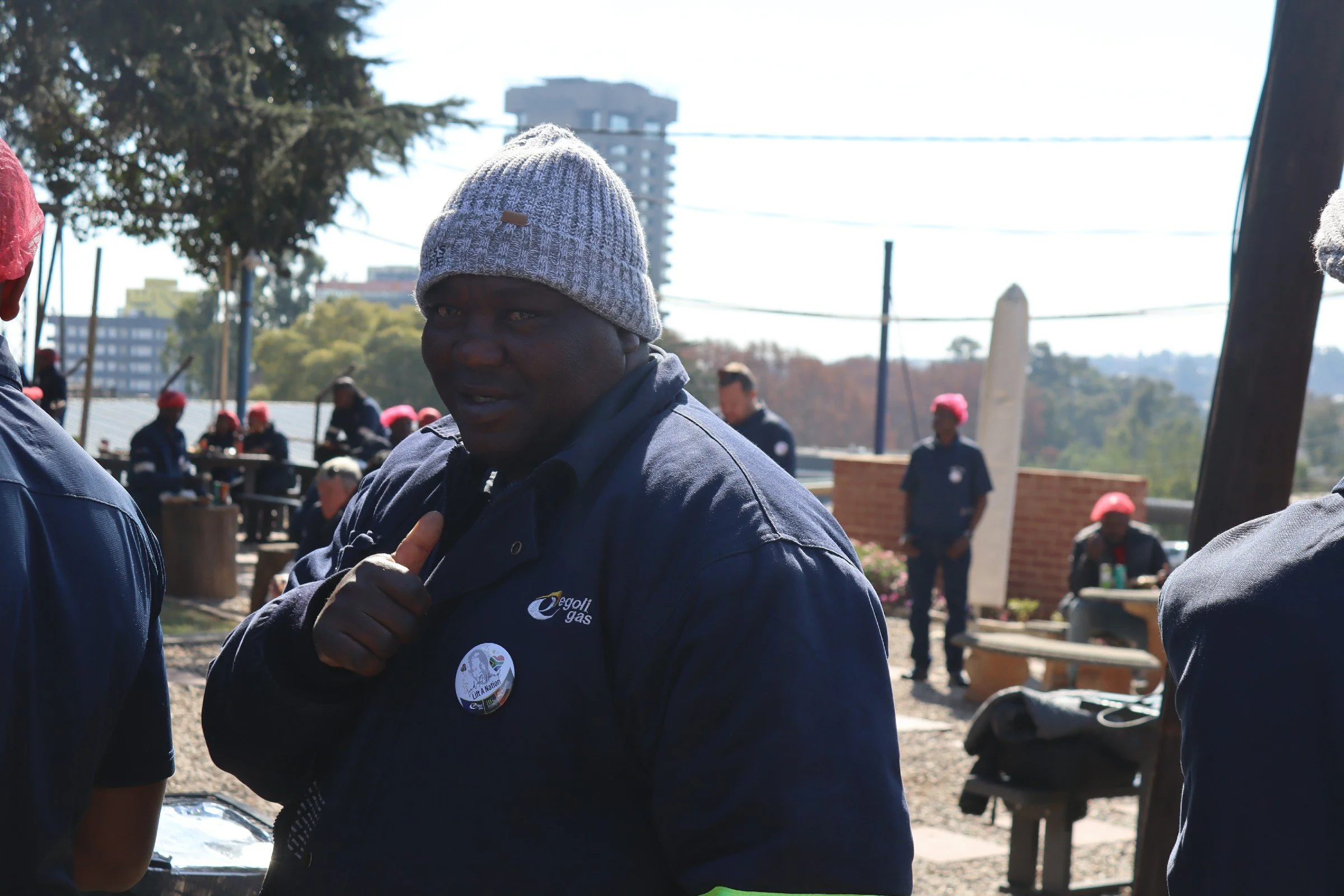 A man wearing a gray knit beanie and a dark blue jacket with a logo on the chest is seen outdoors, talking to someone. There are several people in the background, some wearing red hats, and trees, buildings, and power lines are visible in the distanc