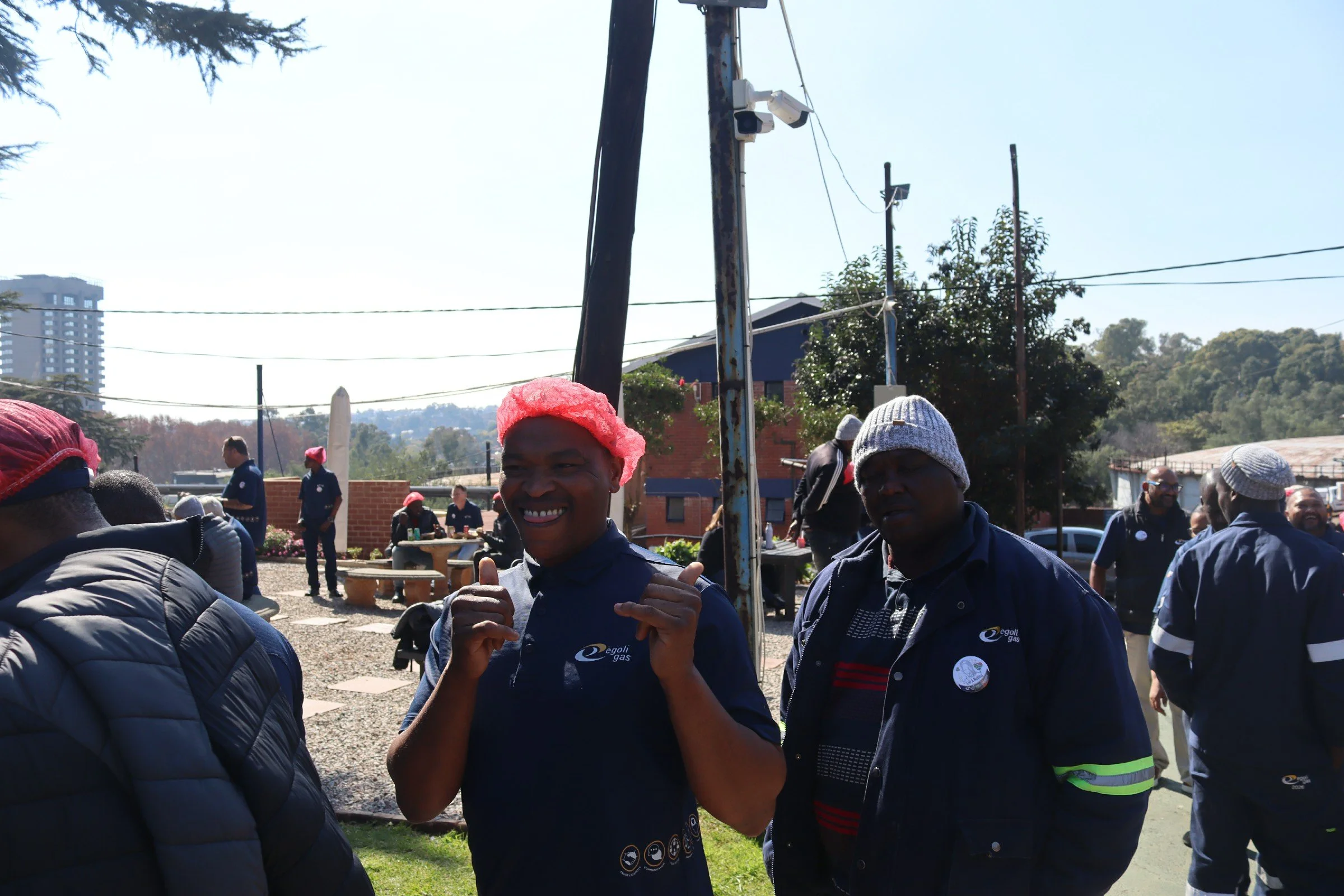 A group of people gathered outdoors on a sunny day, some wearing hats, with a man in the center smiling and giving a thumbs-up. There are trees, buildings, and power lines in the background.