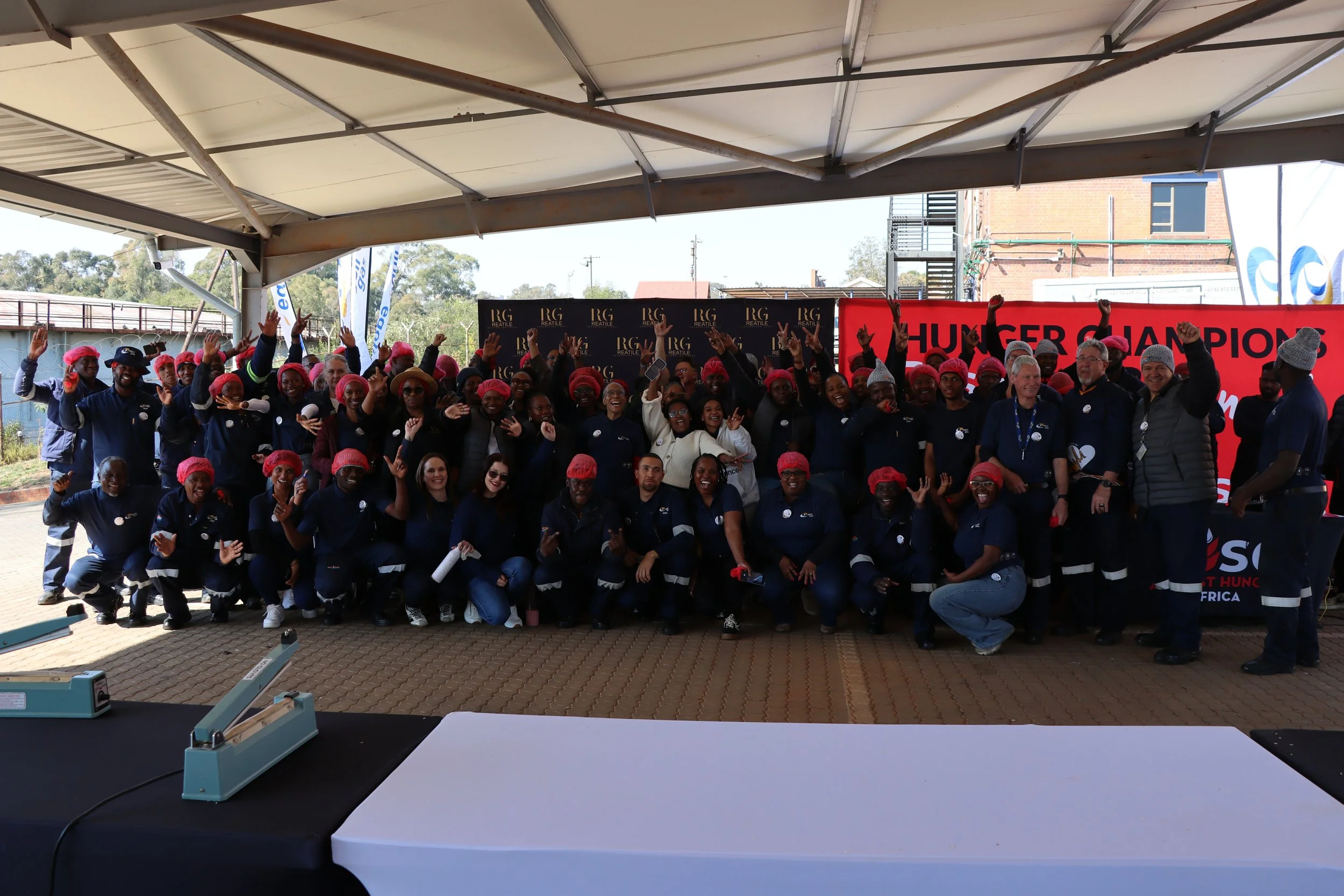 Group of people celebrating a championship victory at an outdoor event, some wearing pink hairnets, standing under a large canopy, with a red banner and black backdrop behind them.