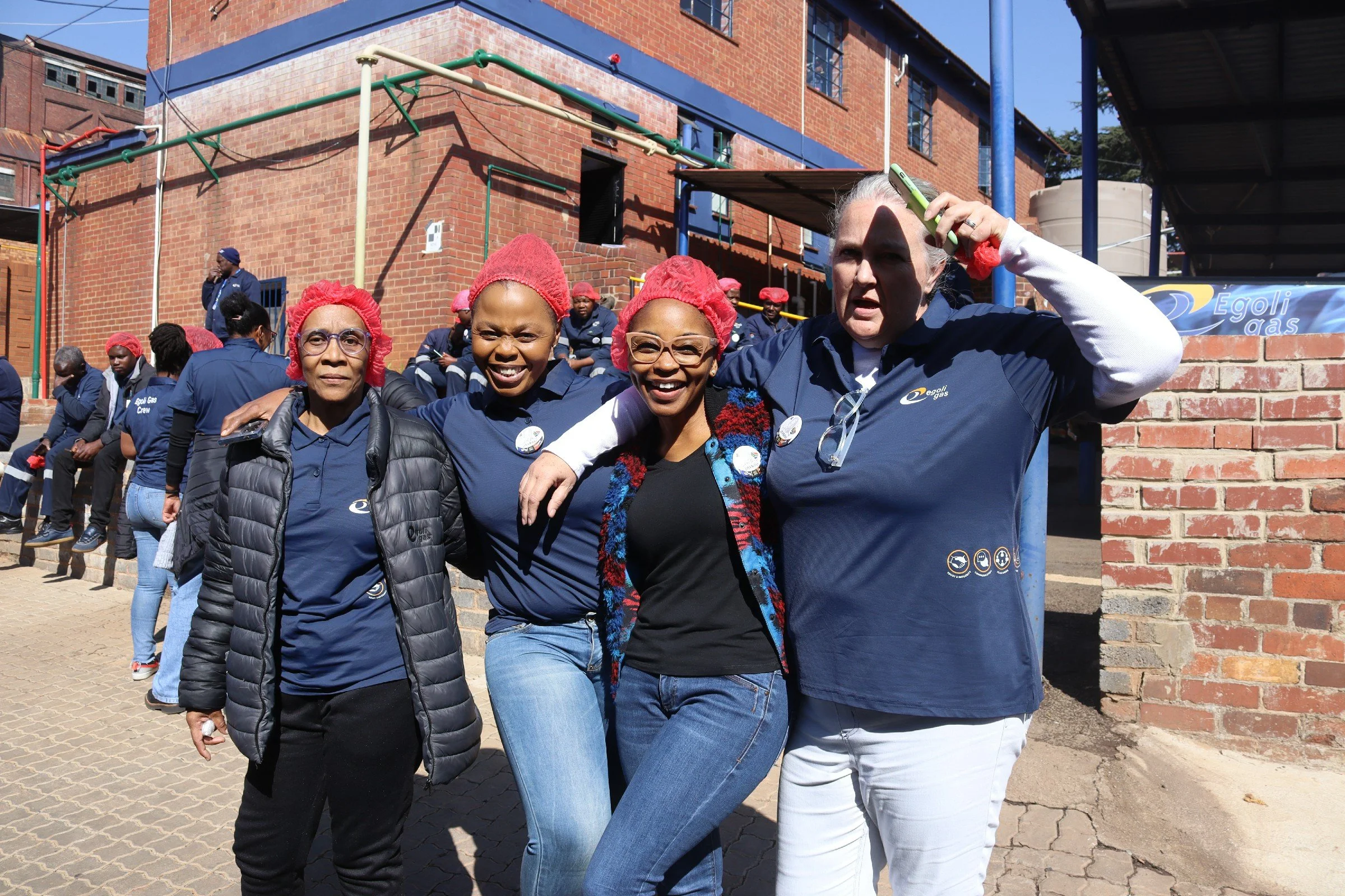 A group of people, mostly women, standing outdoors in front of a brick building. They are wearing blue uniforms with badges and red hairnets. Some are sitting on a ledge, while others stand or lean against a brick wall. The woman in front is smiling,