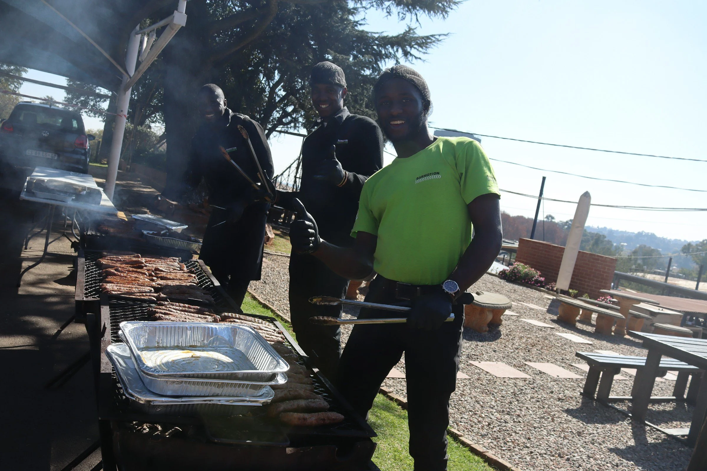 Three young men are standing near a barbecue grill outdoors, cooking meat. They are smiling, giving thumbs up, and appear to be enjoying a cookout on a sunny day with trees and a parking area in the background.