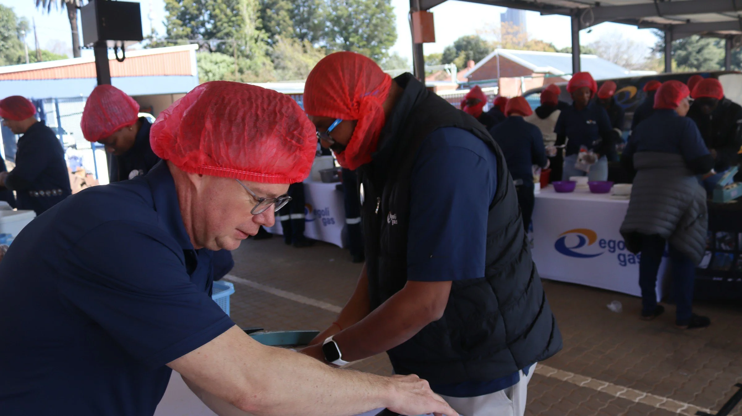 Two people wearing red hairnets and dark clothing working together at an indoor event, with a table displaying a sign for "Oregon Gas" in the background.