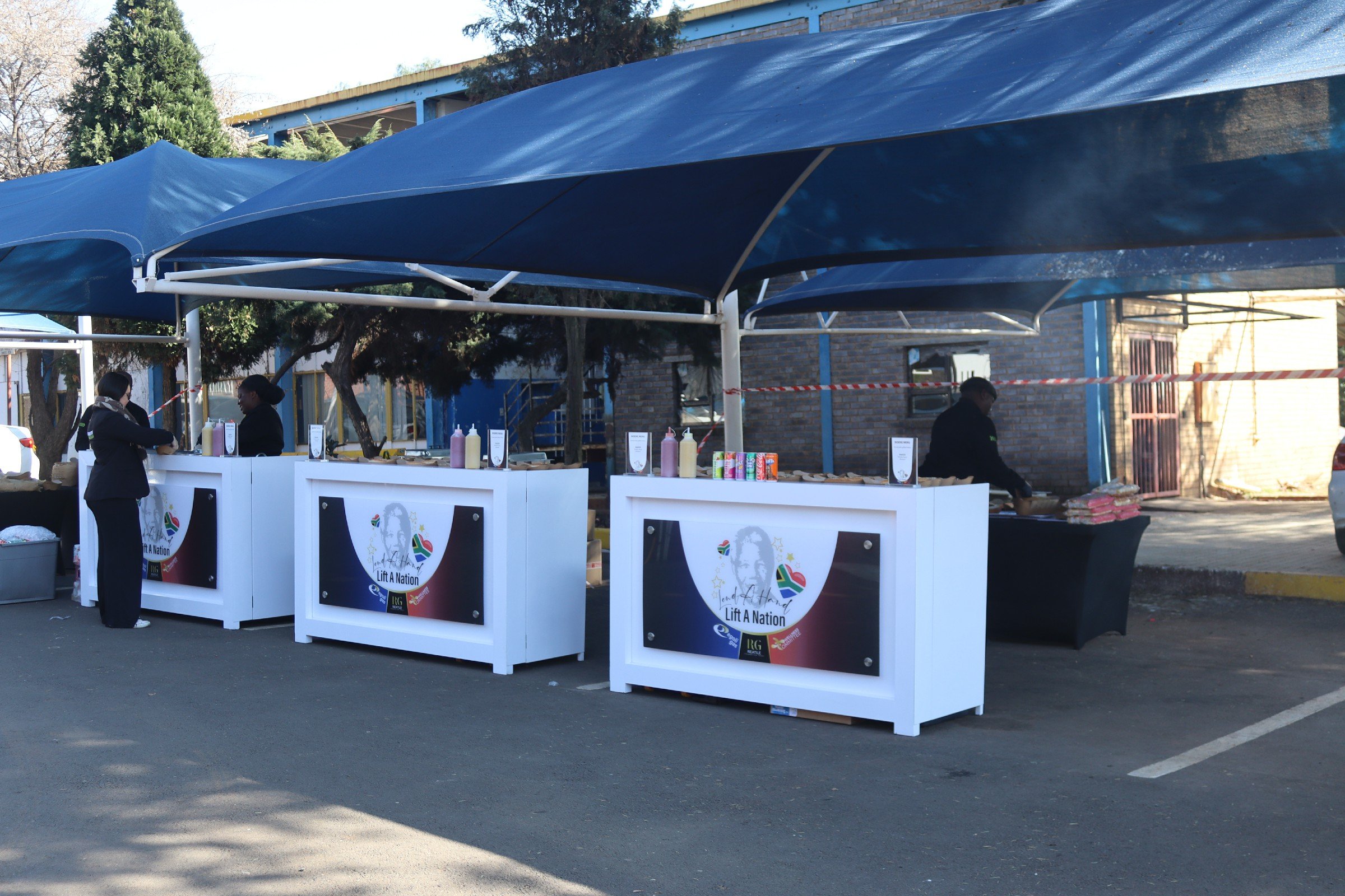 Outdoor market stalls with blue canopies and signs that say 'Lift A Nation,' with people working at the stalls against a background of trees and buildings.