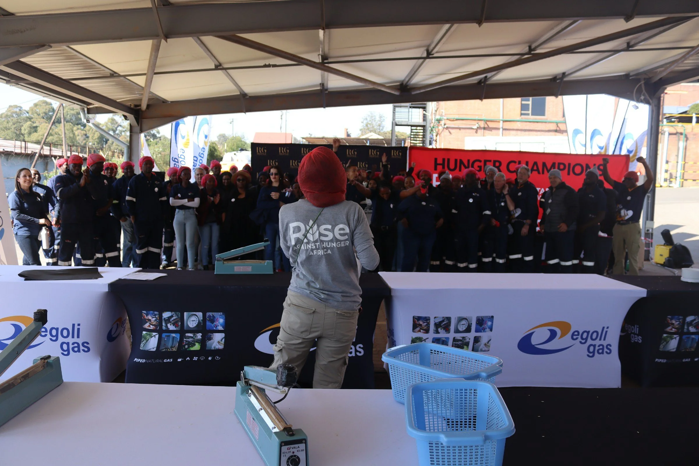 A group of people wearing matching dark uniforms and pink head coverings gathered under a large outdoor tent, participating in an event related to hunger awareness. A woman with long dark hair in sunglasses stands in front of the group, speaking or l