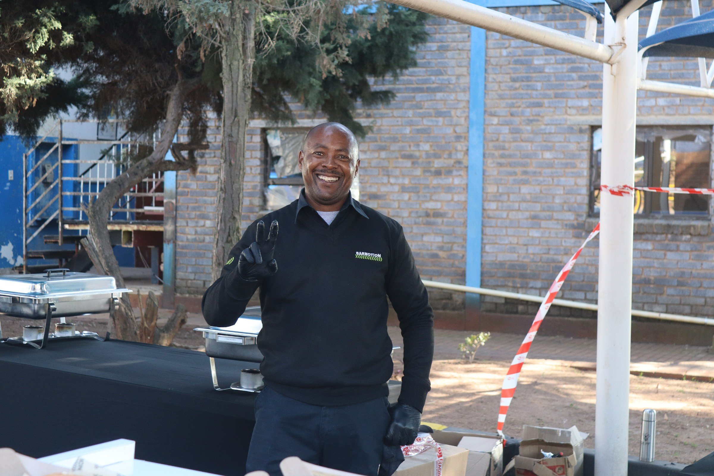 A smiling man wearing a black jacket and gloves making a peace sign with his right hand at an outdoor event, with food trays and tables in front of him, trees, and a brick building in the background.