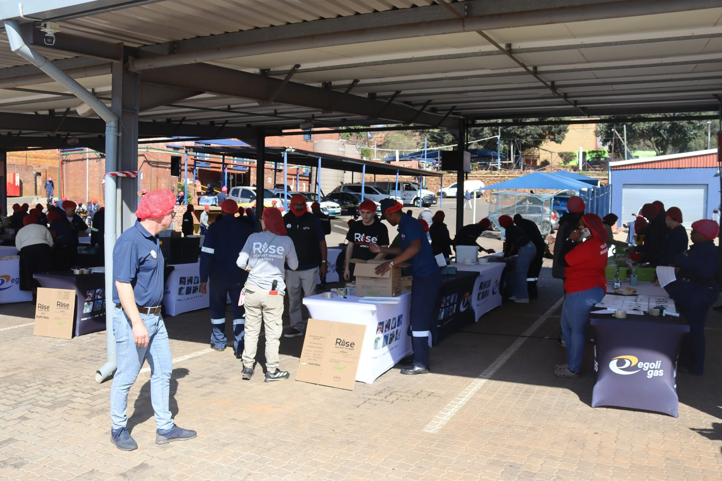 People wearing red hairnets and dark blue uniforms gather under a covered outdoor area, engaging in an event or activity, with tables and boxes present. Vehicles are parked in the background, and signage includes 'Rise Against Hunger' and 'Egoli Gas'