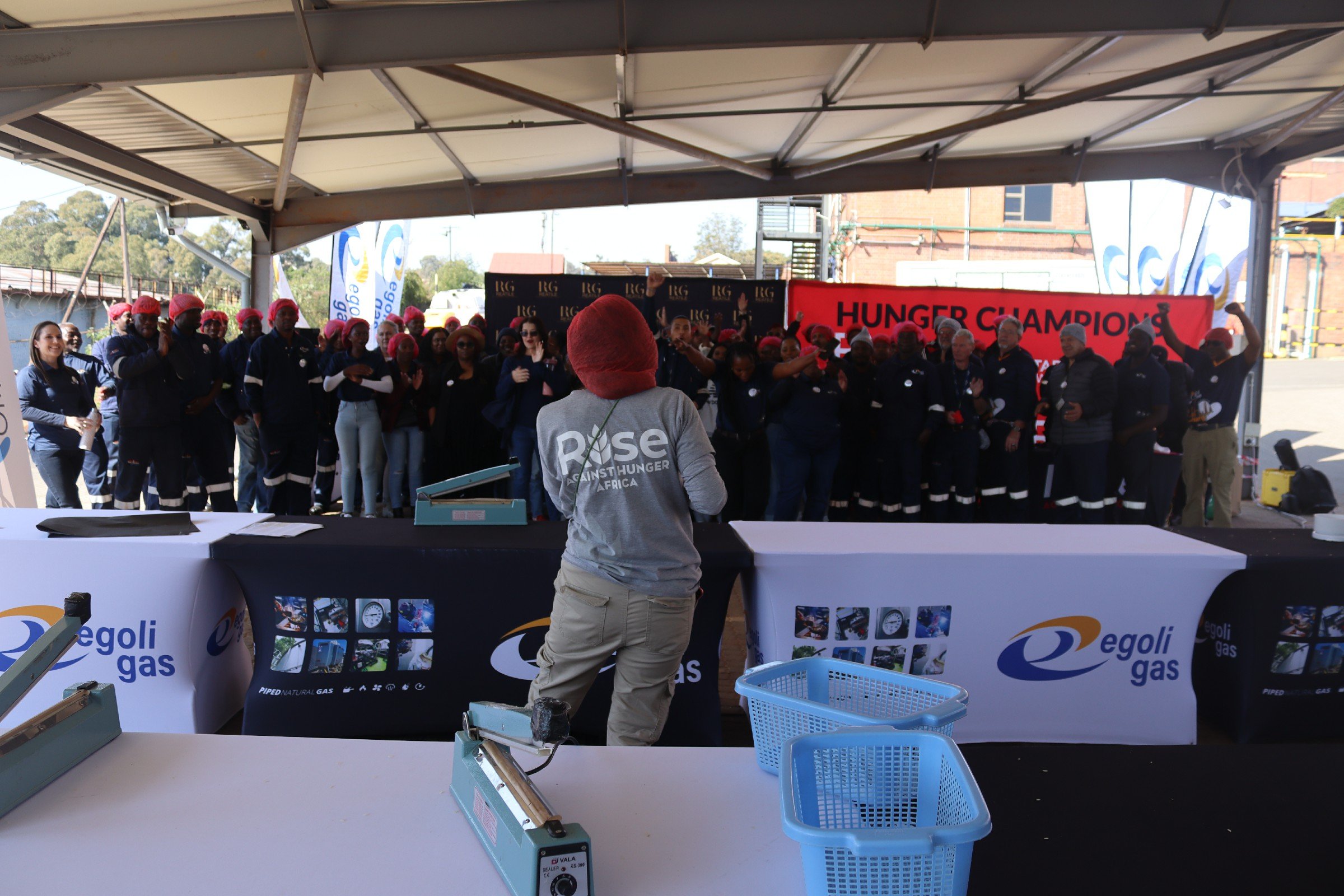 A large group of people, many wearing red head coverings, gathered under a tent at a public event. A woman with sunglasses is giving a speech, and there are banners that read 'Hunger Champions.' The setting appears to be outdoors in an urban or indus