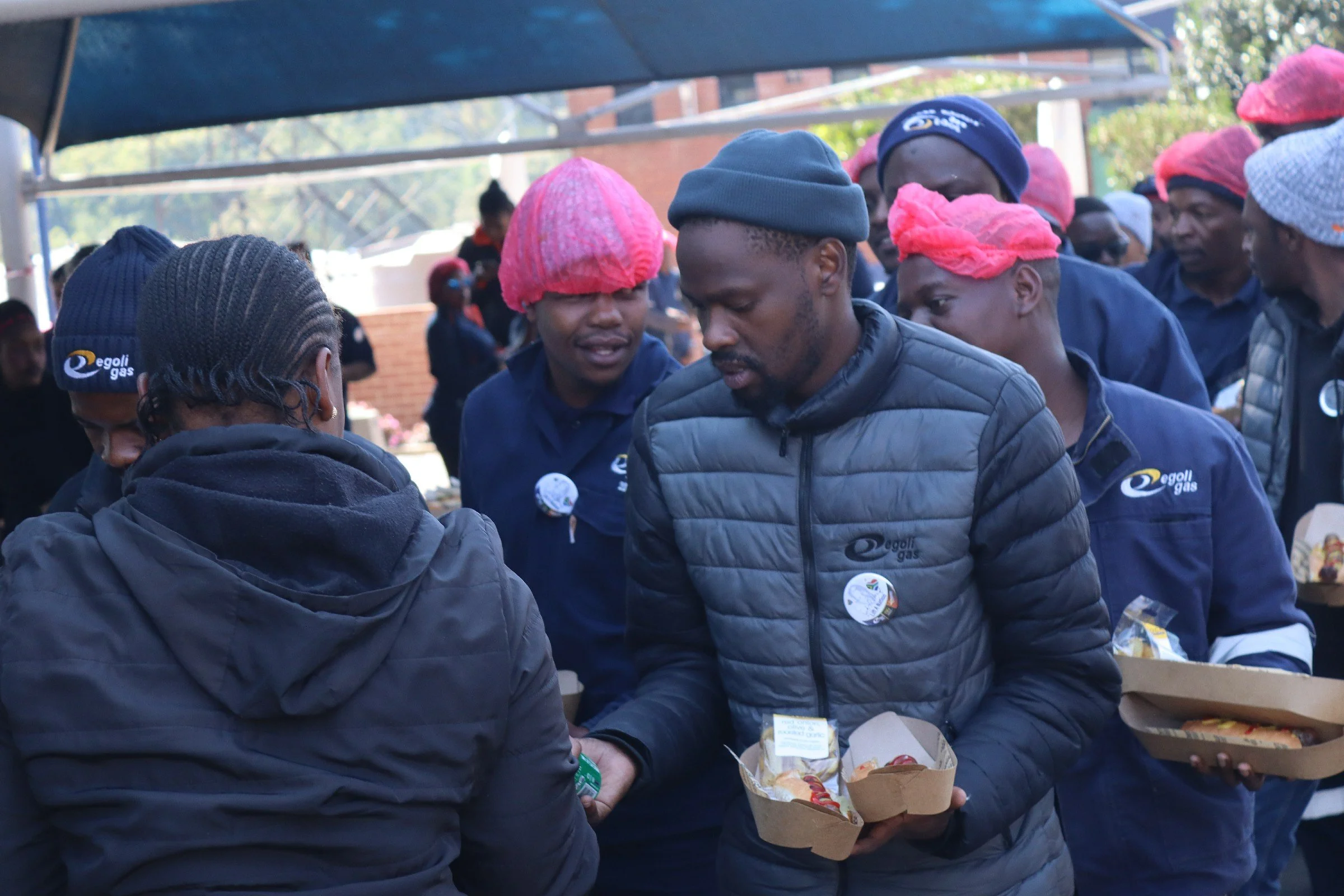 People gathered at an outdoor event, wearing jackets and colorful hair coverings, handing out food and drinks on a sunny day.