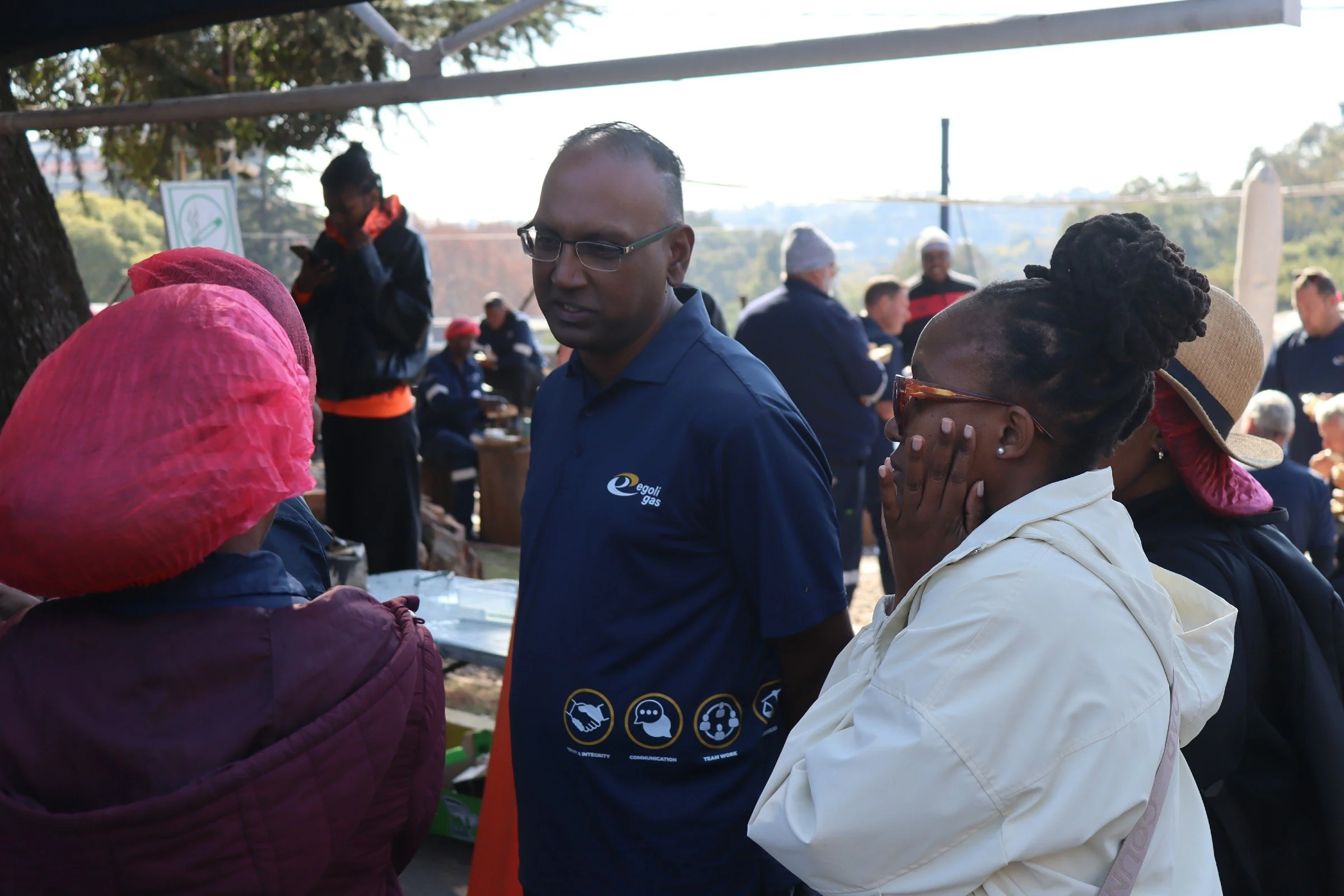 People gathered outdoors at a daytime event, some wearing jackets and head coverings, with a man in glasses and a blue polo shirt in the center, surrounded by women in headscarves and a woman with dreadlocks and sunglasses, engaged in conversation.