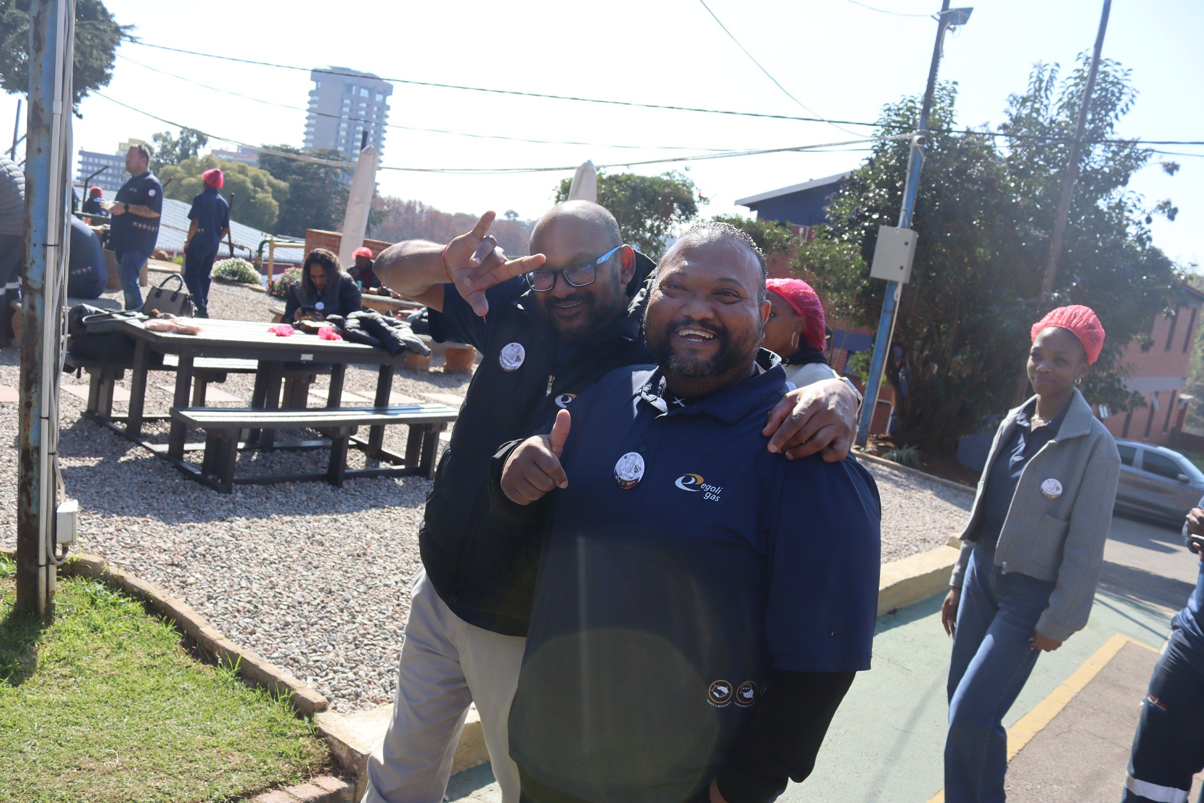 Two men smiling and posing for a photo outdoors, one making a peace sign, with several other people in the background at a public gathering or event.