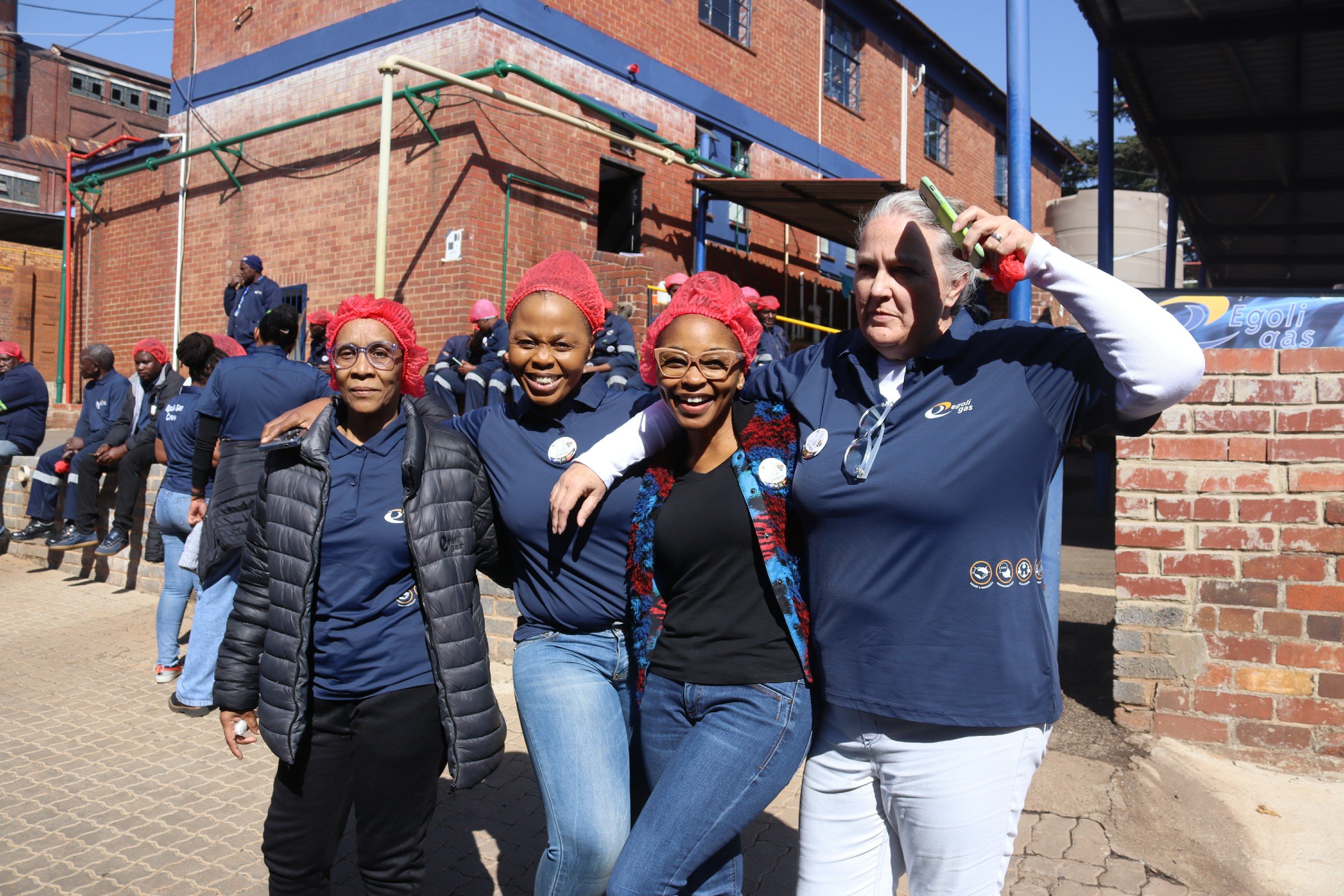 Group of women and girls standing outdoors in front of a brick building, smiling and posing for the photo. Some wear red hairnets and jackets with the Egoli Gas logo, indicating participation in a community event or gathering.