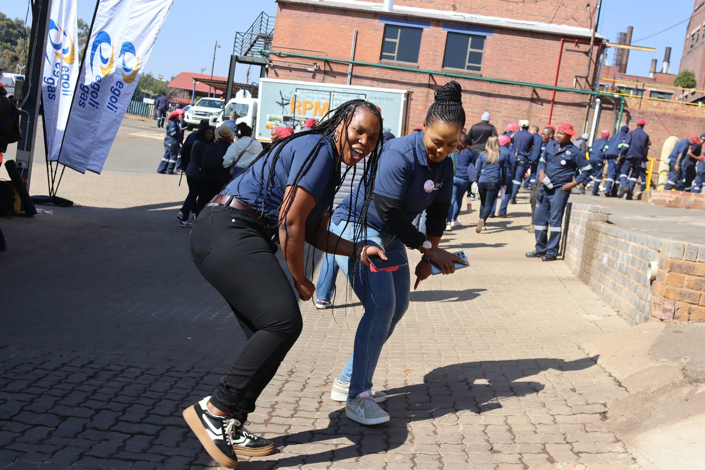 Two women with long braided hair are dancing or celebrating outdoors on a brick paved area, with one wearing black sneakers and the other wearing white sneakers, amidst a crowd of people and flags with the logo 'Galil' and the text 'Royal.' The crowd
