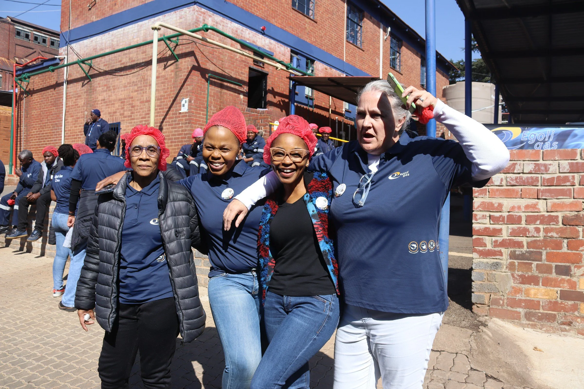 Group of people, mostly women, standing outdoors in front of a brick building, wearing blue shirts and red hair coverings, some smiling and some looking serious.