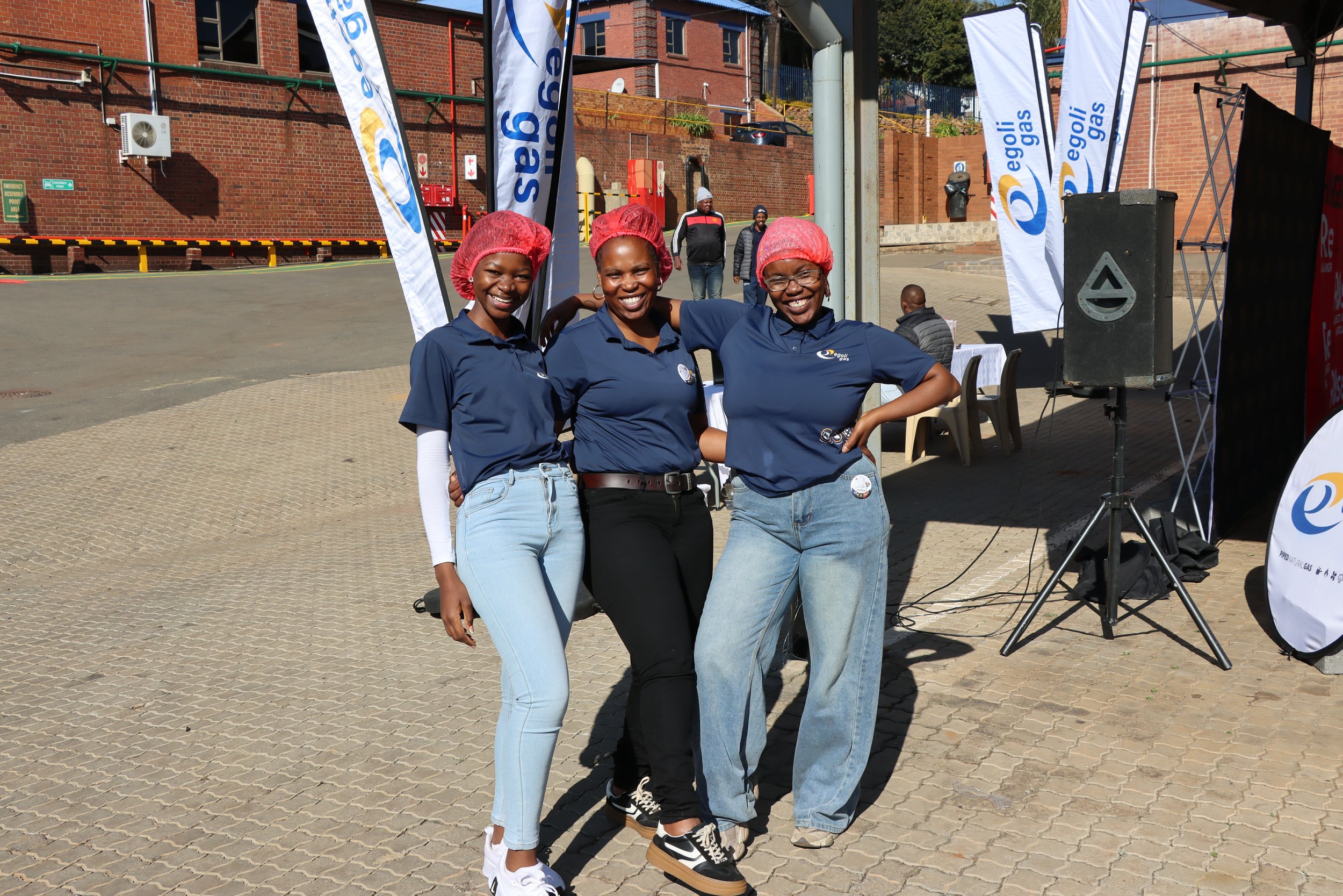 Three women wearing blue shirts and red hairnets smiling and posing together outdoors near a setup with banners and a speaker, with a brick building in the background.