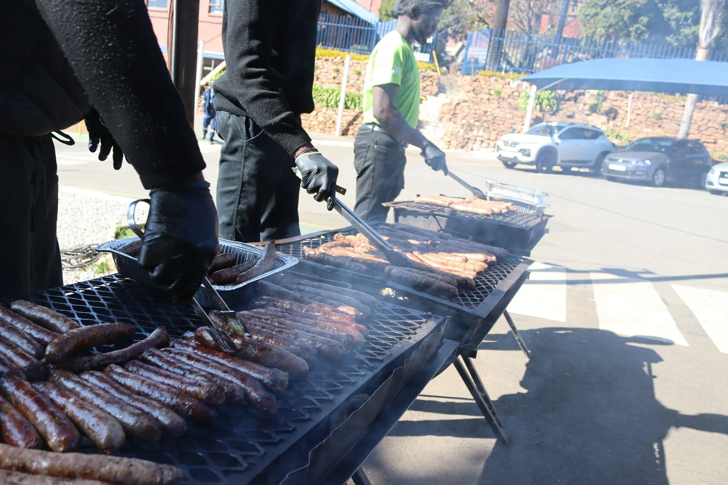 A group of people cooking and grilling sausages and meat on outdoor barbecue grills during daytime.