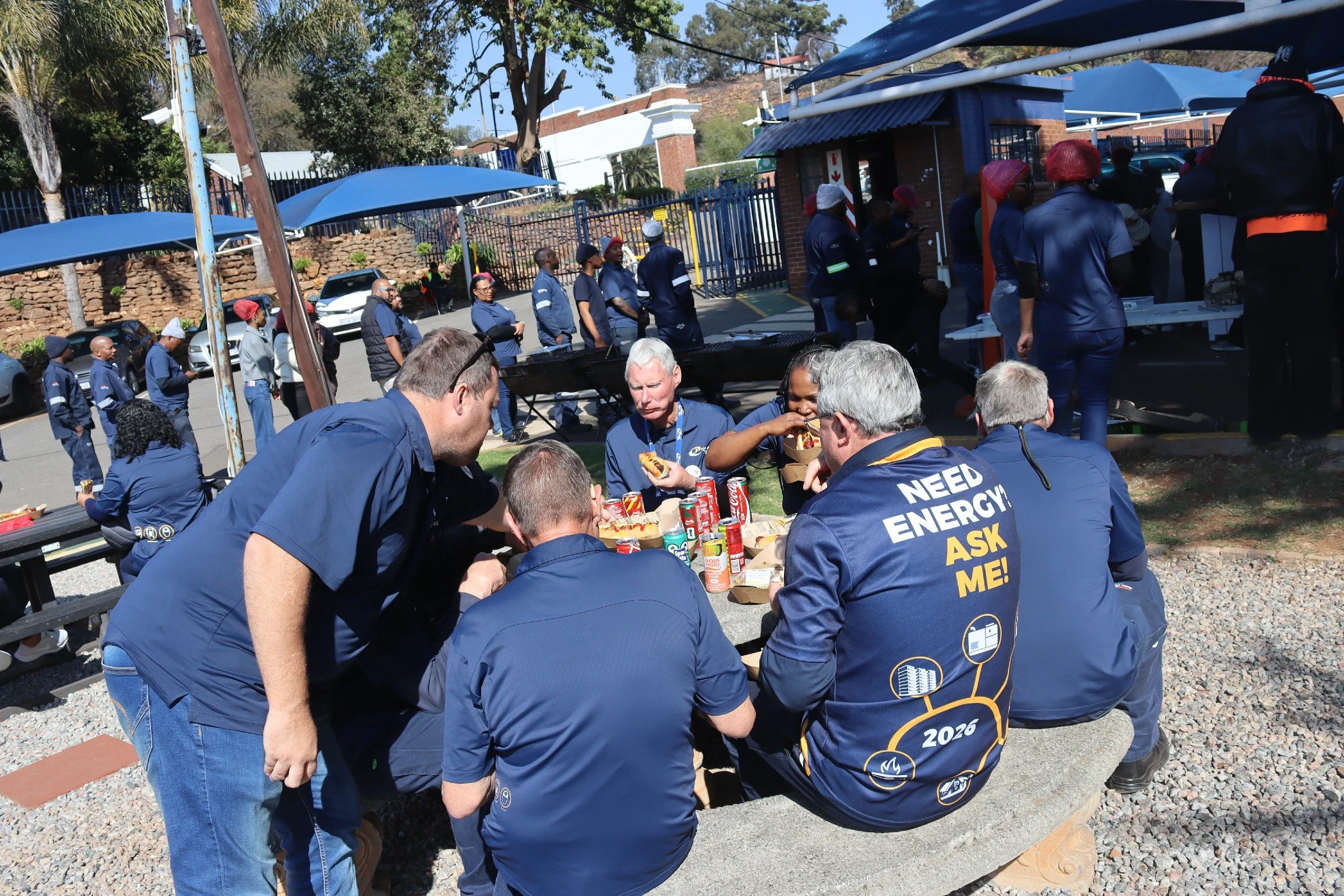 A group of people, possibly workers or volunteers, gathered around a table outdoors, eating and drinking, with some wearing navy shirts with safety or organizational logos. In the background, more people are standing in line near a fenced area with b