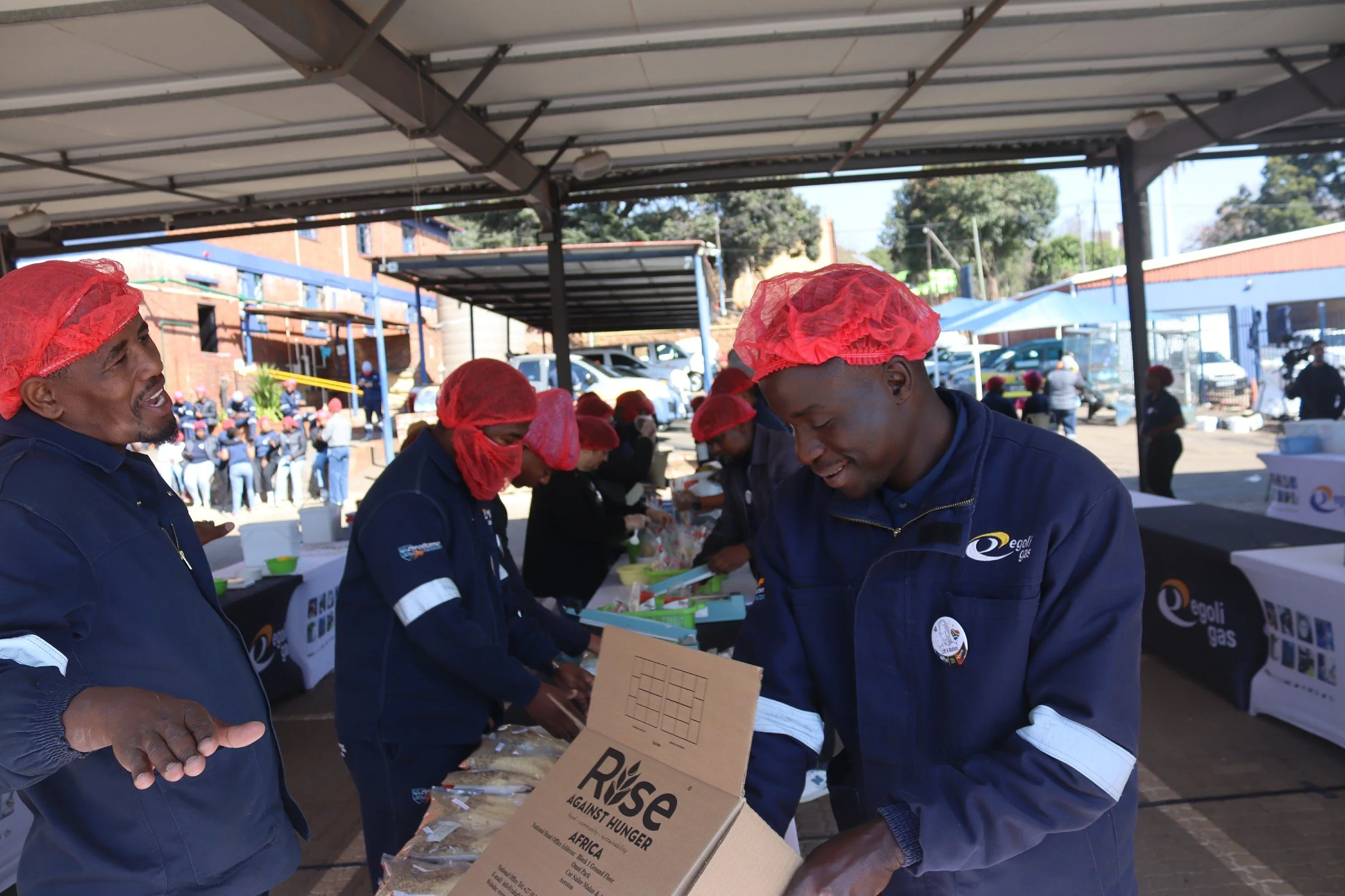 A group of people wearing blue uniforms and red hair covers participating in an event, with some holding boxes and banners, outside under a large canopy with parked cars and buildings in the background.