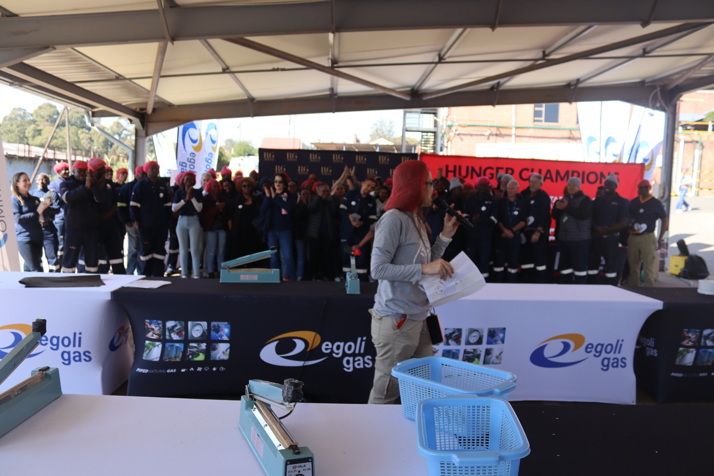 A group of people, many wearing red hats, gathered under a tent at an outdoor event for a Hunger Challenge. A woman with pink hair is speaking into a microphone, and there are banners and signs related to Egoli Gas in the background, with tables and 