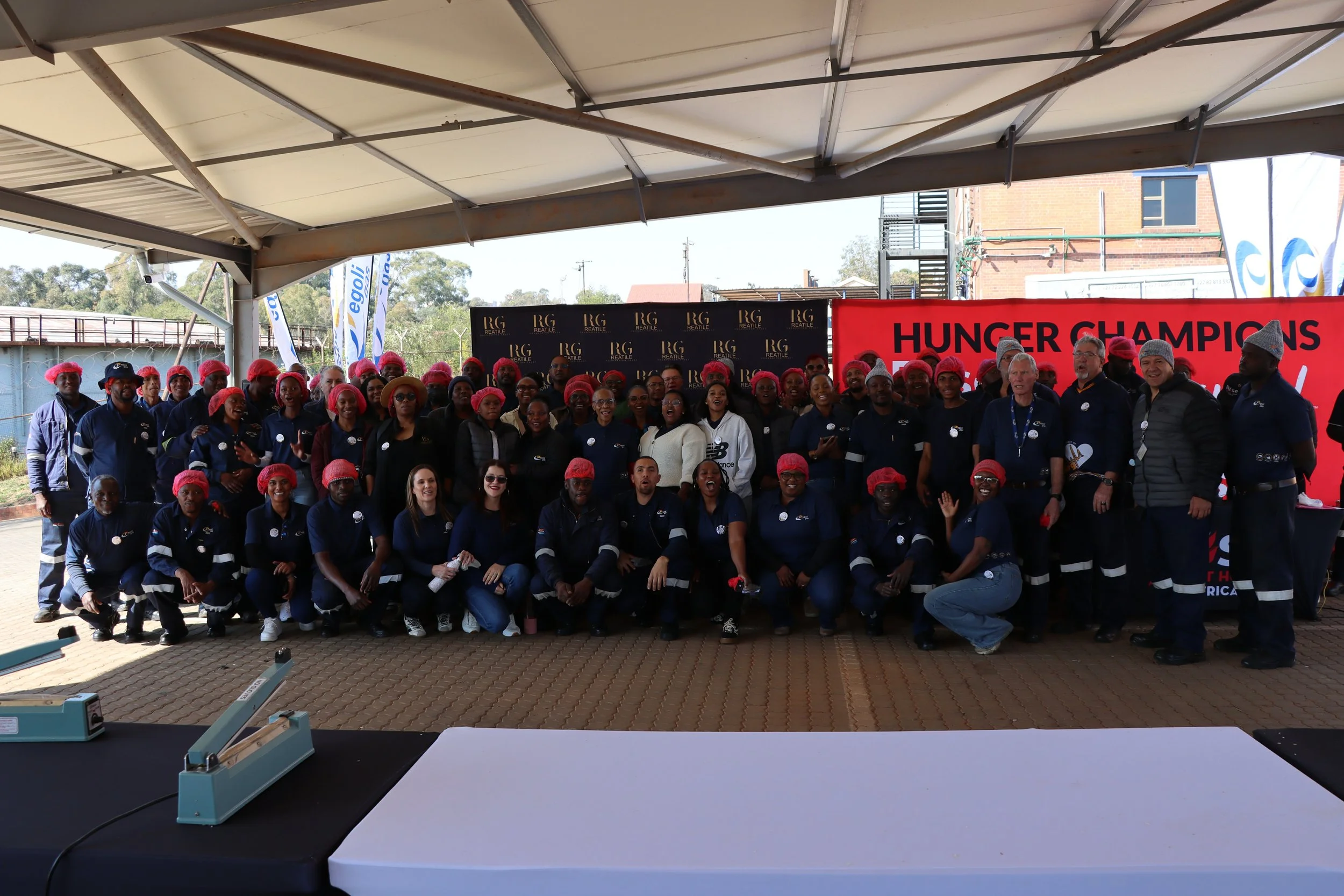 Group of people wearing red hair nets, standing and smiling under a shelter, with banners and a red sign in the background that reads 'Hunger Champions'.