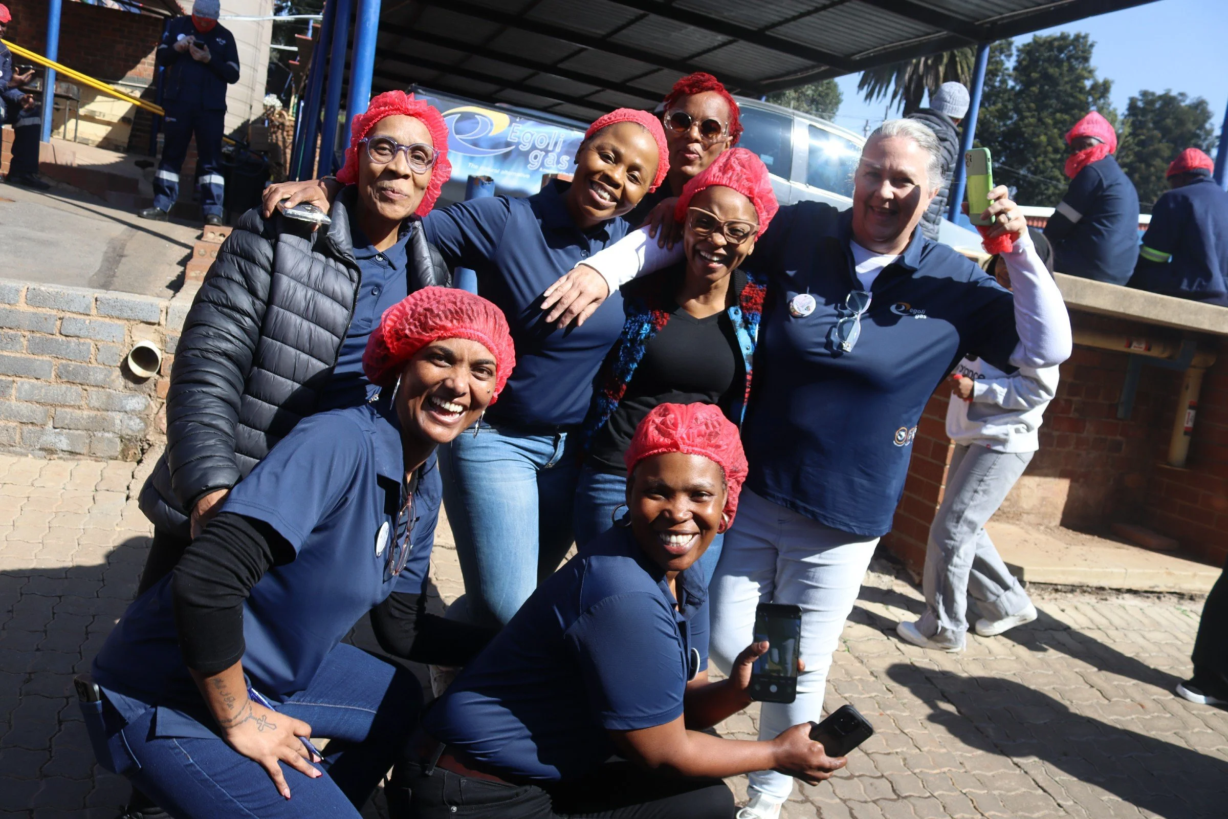Group of people smiling and posing for a photo outdoors, many wearing red hairnets and blue shirts, with some holding phones.