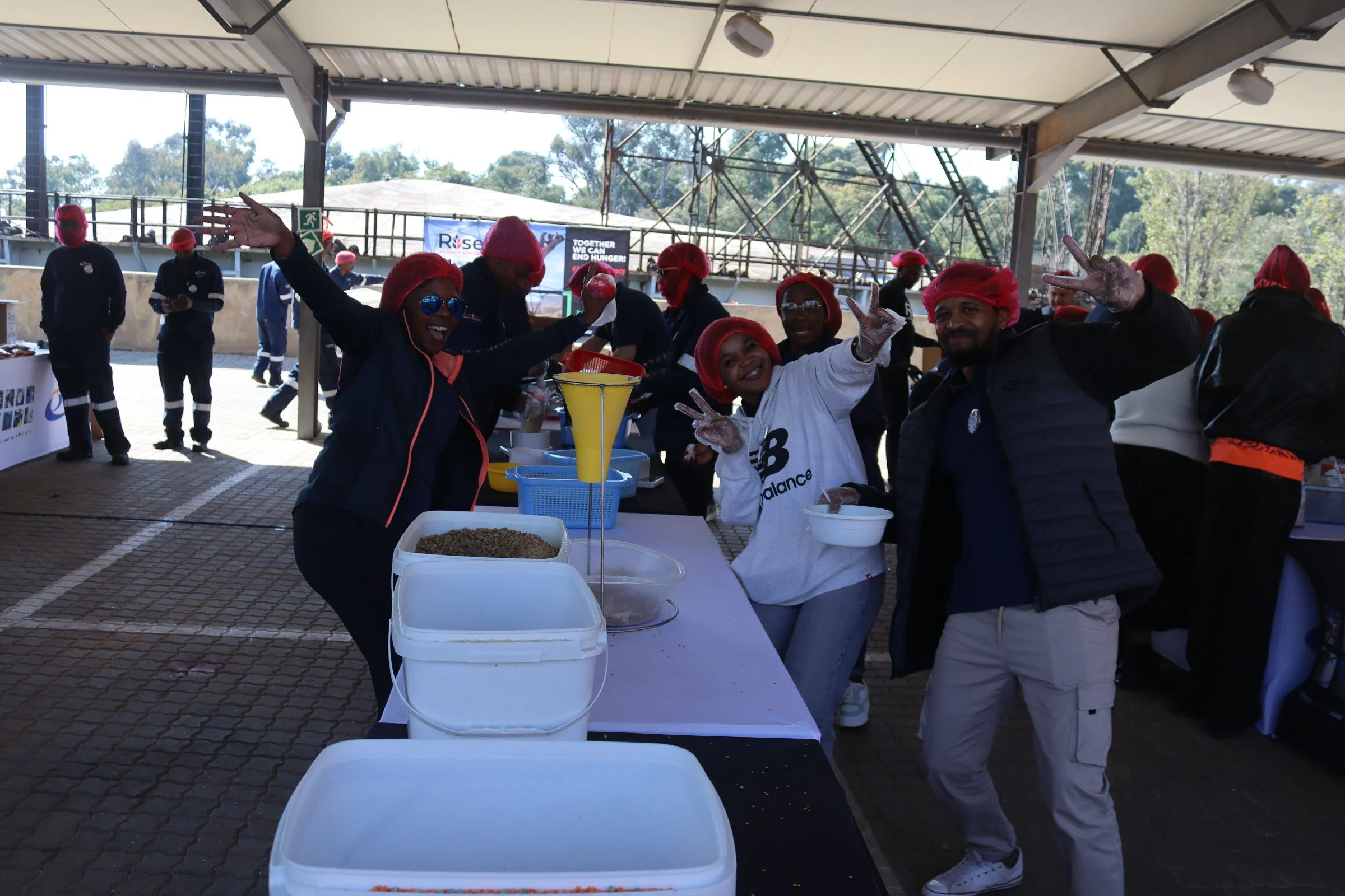 A group of people with red hairnets and gloves, smiling and making peace signs at a food serving event, with tables holding food containers in the foreground and a stainless steel roof overhead.