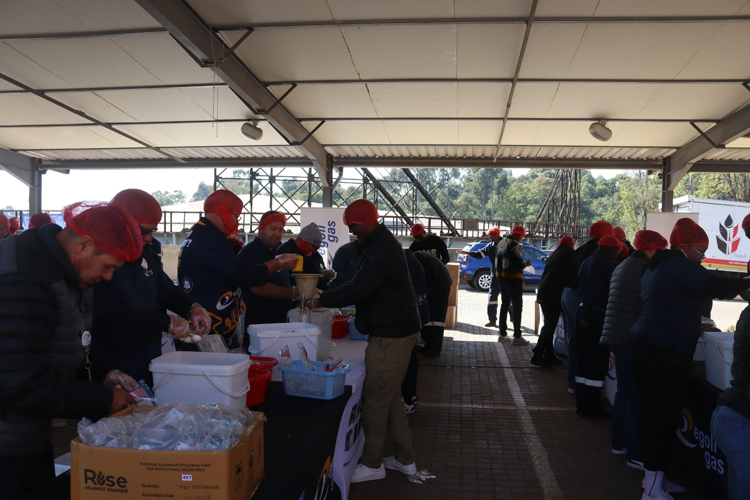 People wearing red hairnets and gloves serving food at an outdoor event with tables, boxes, and a banner that reads 'Edelgas' and 'Gas'.